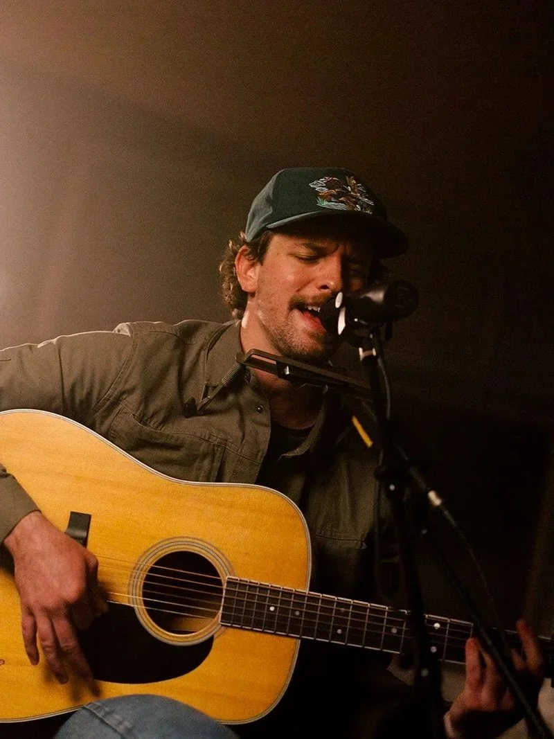 A man with a black baseball cap, brown jacket, and black shirt is singing into a microphone while playing an acoustic guitar.