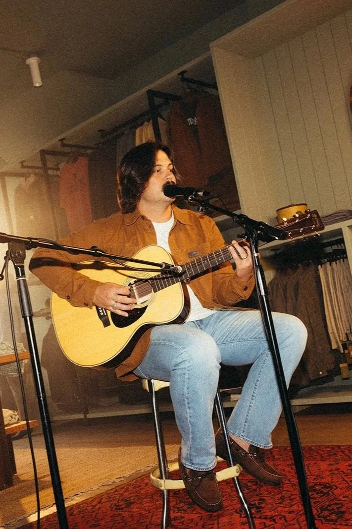 A man with shoulder-length dark hair is sitting on a stool, playing an acoustic guitar and singing into a microphone. He is wearing a brown jacket, white shirt, and light blue jeans. The background shows a cozy room with wooden paneling, some hanging