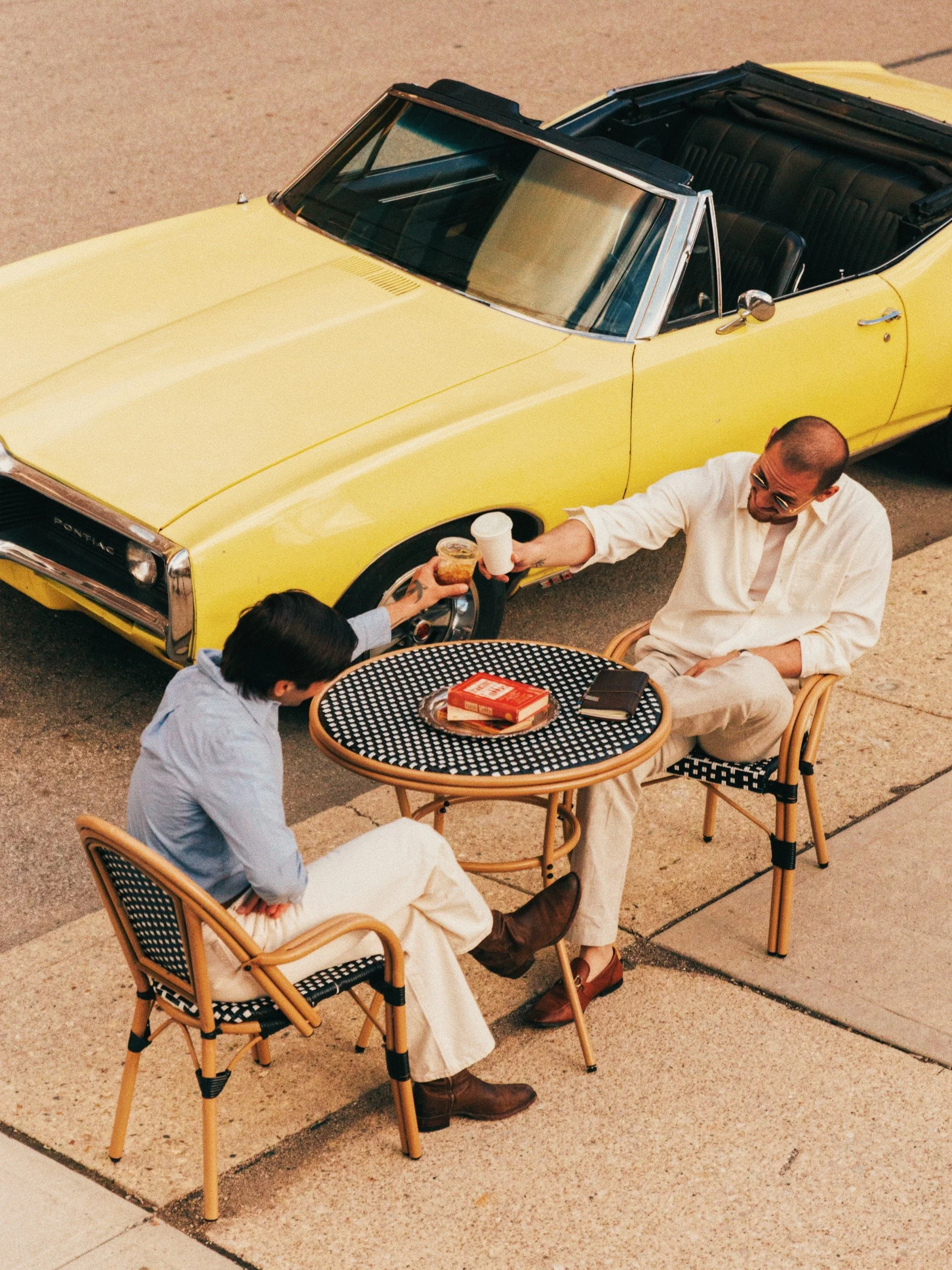 Two men are sitting at a black and white checkered outdoor table on a sidewalk, having drinks. One man in a beige outfit and sunglasses is smiling, while the other man, wearing a light blue shirt, is reaching for a drink. Cafe Stai Manso