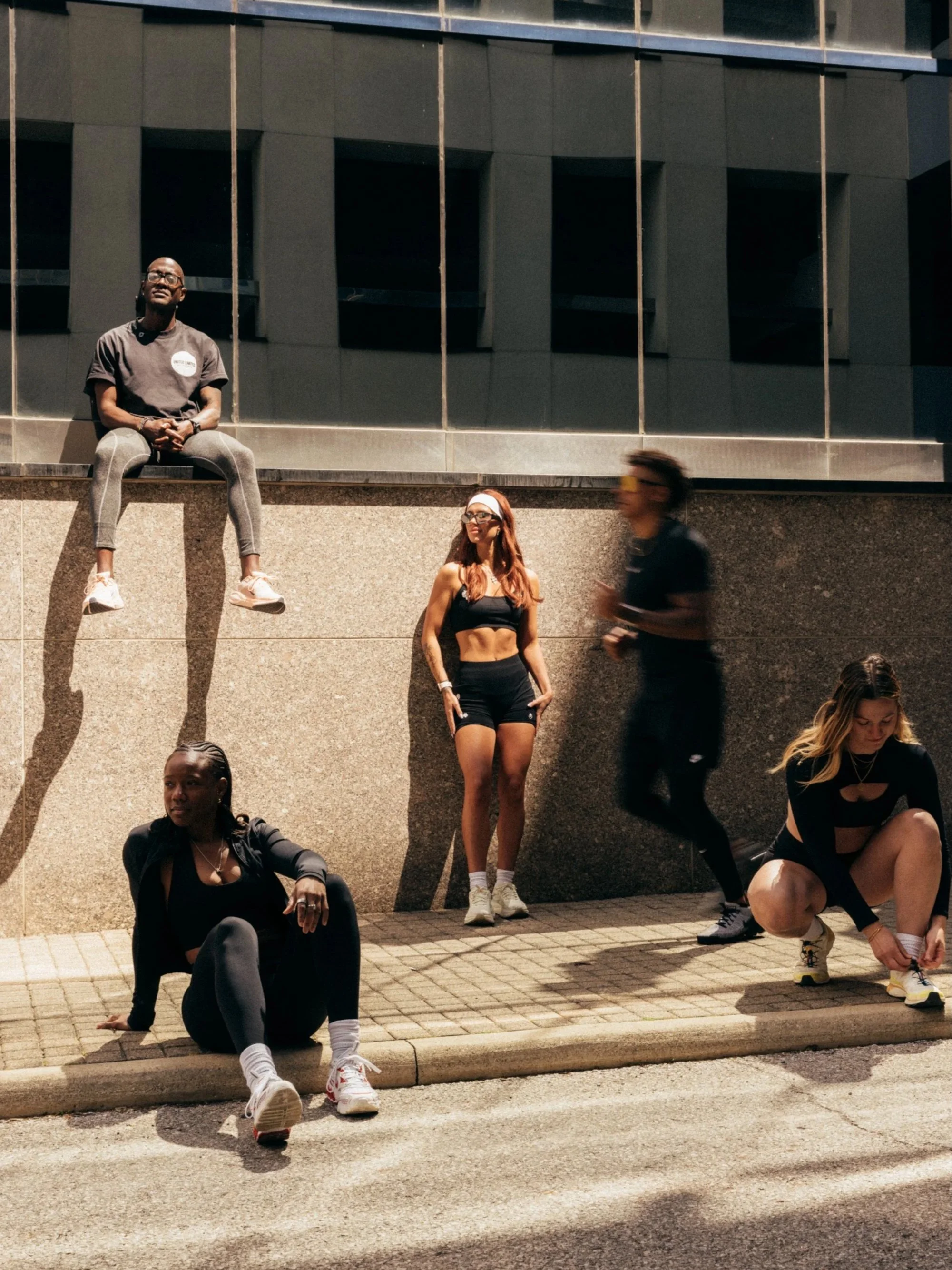 Five young people posing casually outside near a building with large rectangular windows. Some are sitting or squatting on the ground, one is sitting on a ledge, and one is standing. They wear athletic or casual clothing, and two of them are wearing 