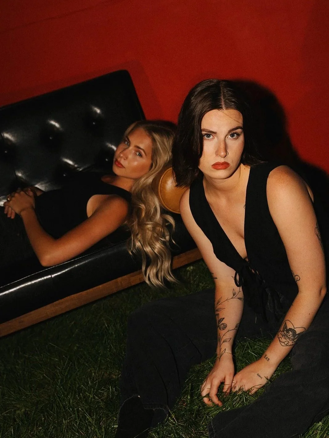 Two women, one with long blonde hair lying on a black leather couch, and the other with short dark hair sitting on grass in front of the couch, both wearing black outfits. The background is red.