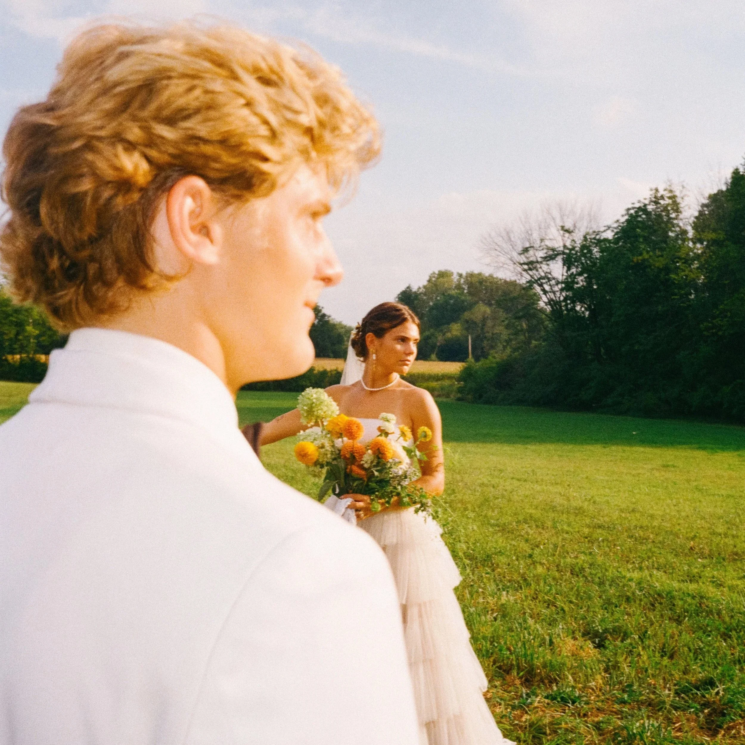 A wedding scene with a man in a white suit in the foreground and a bride holding a bouquet of flowers in the background, outdoors on a grassy field under a partly cloudy sky.