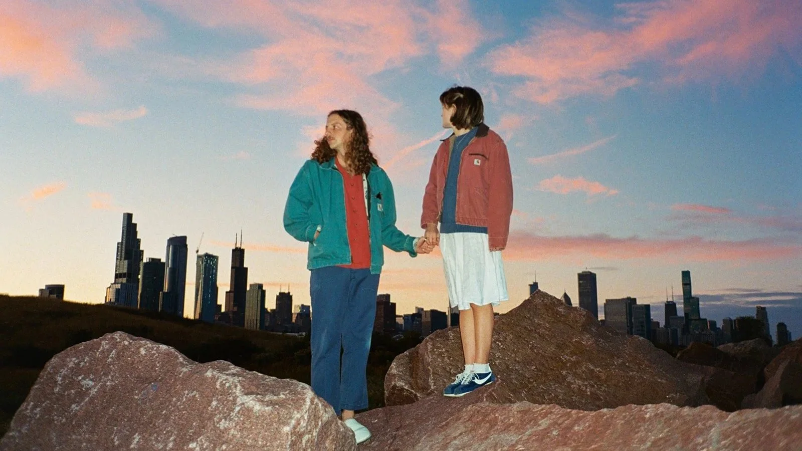 Two people holding hands on large rocks during sunset with the Chicago skyline in the background.
