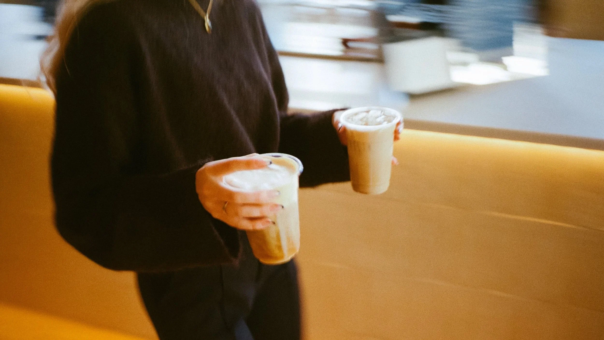 A person holding two iced coffee drinks inside a cafe with a yellow bench and a window in the background.