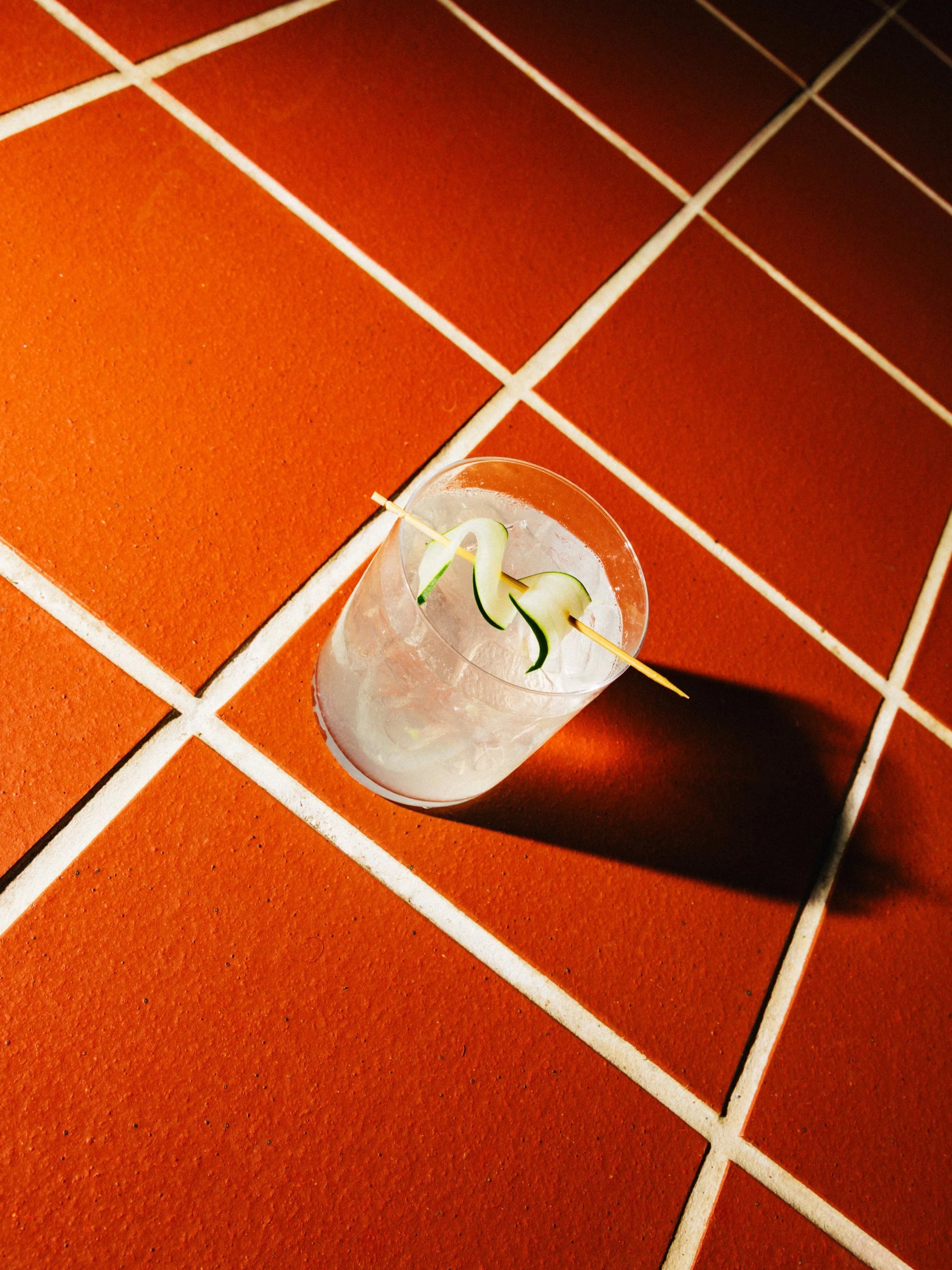 A clear glass with a cocktail garnished with a thin slice of cucumber, placed on a terracotta tiled floor.