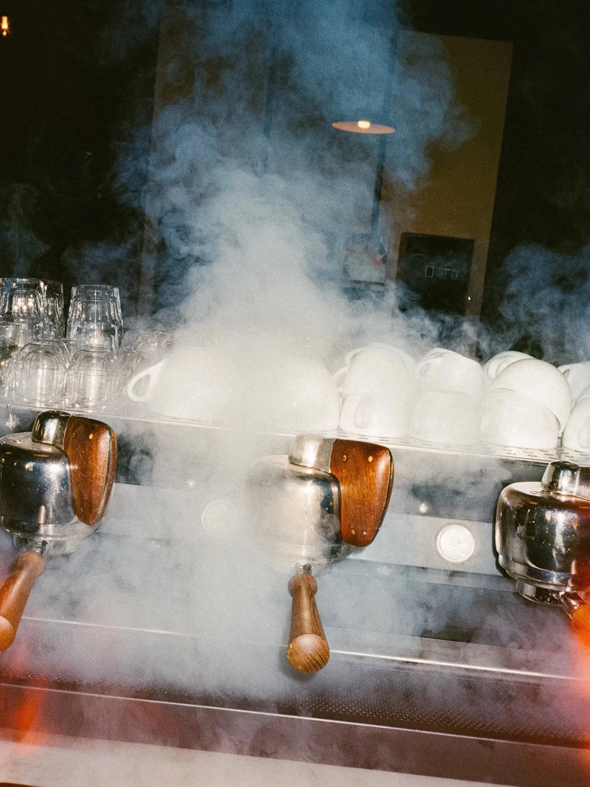 An espresso machine with steam rising from it, surrounded by several white cups and glasses.