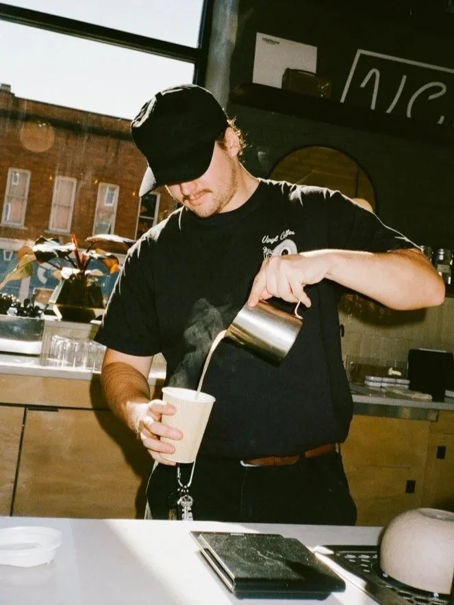 A barista pouring steamed milk into a paper cup at a coffee shop with a large window behind him