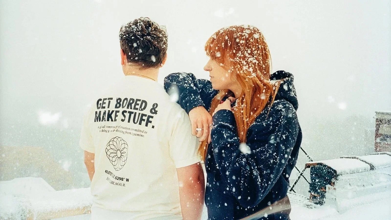 Two people stand close together in a snowy outdoor setting, one with their back to the camera wearing a white shirt with bold text, the other facing sideways with long red hair and a dark jacket, snow falling around them.