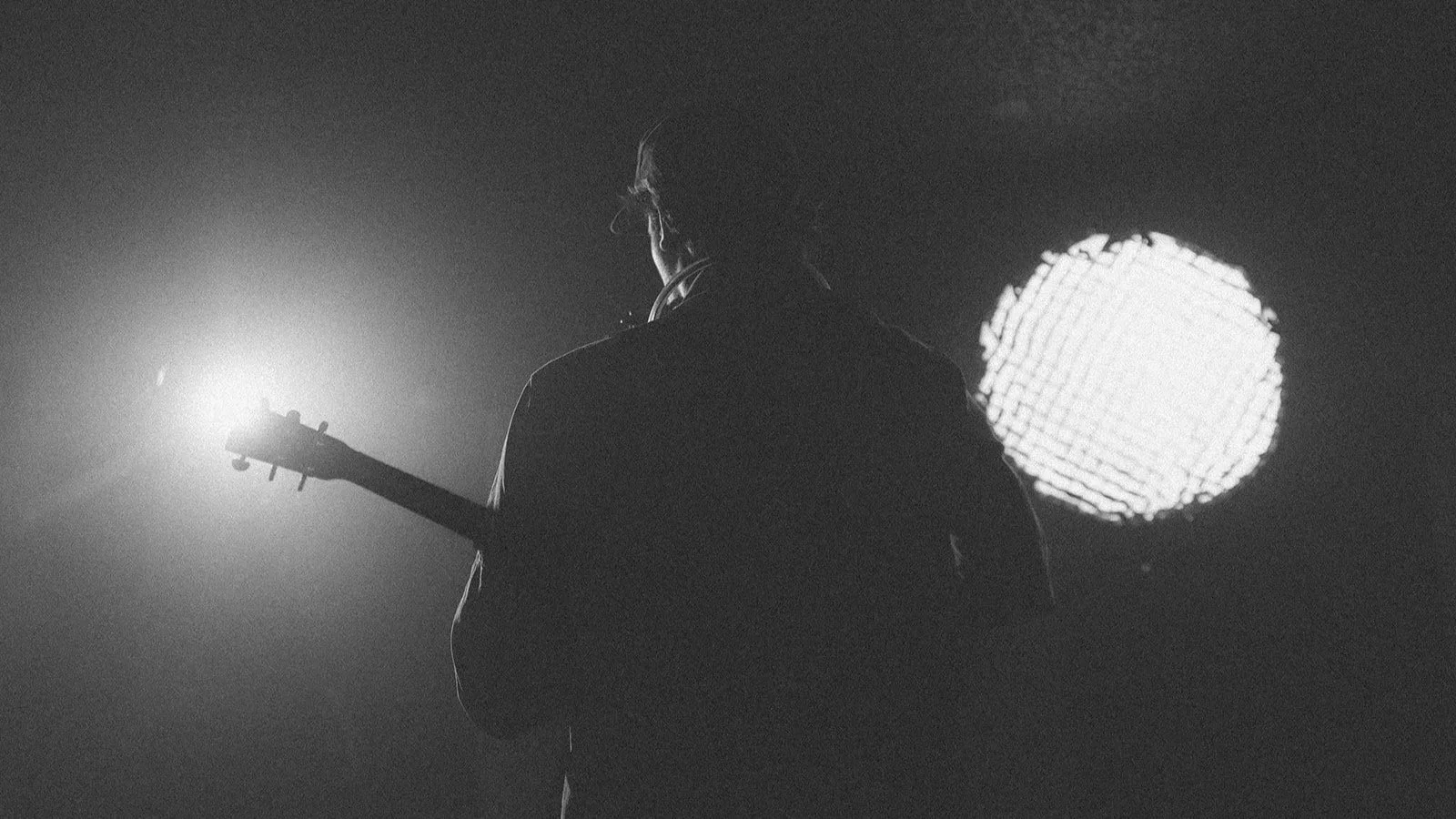 A silhouette of a person holding a guitar in front of bright stage lights, with one bright light behind them creating a contrast.