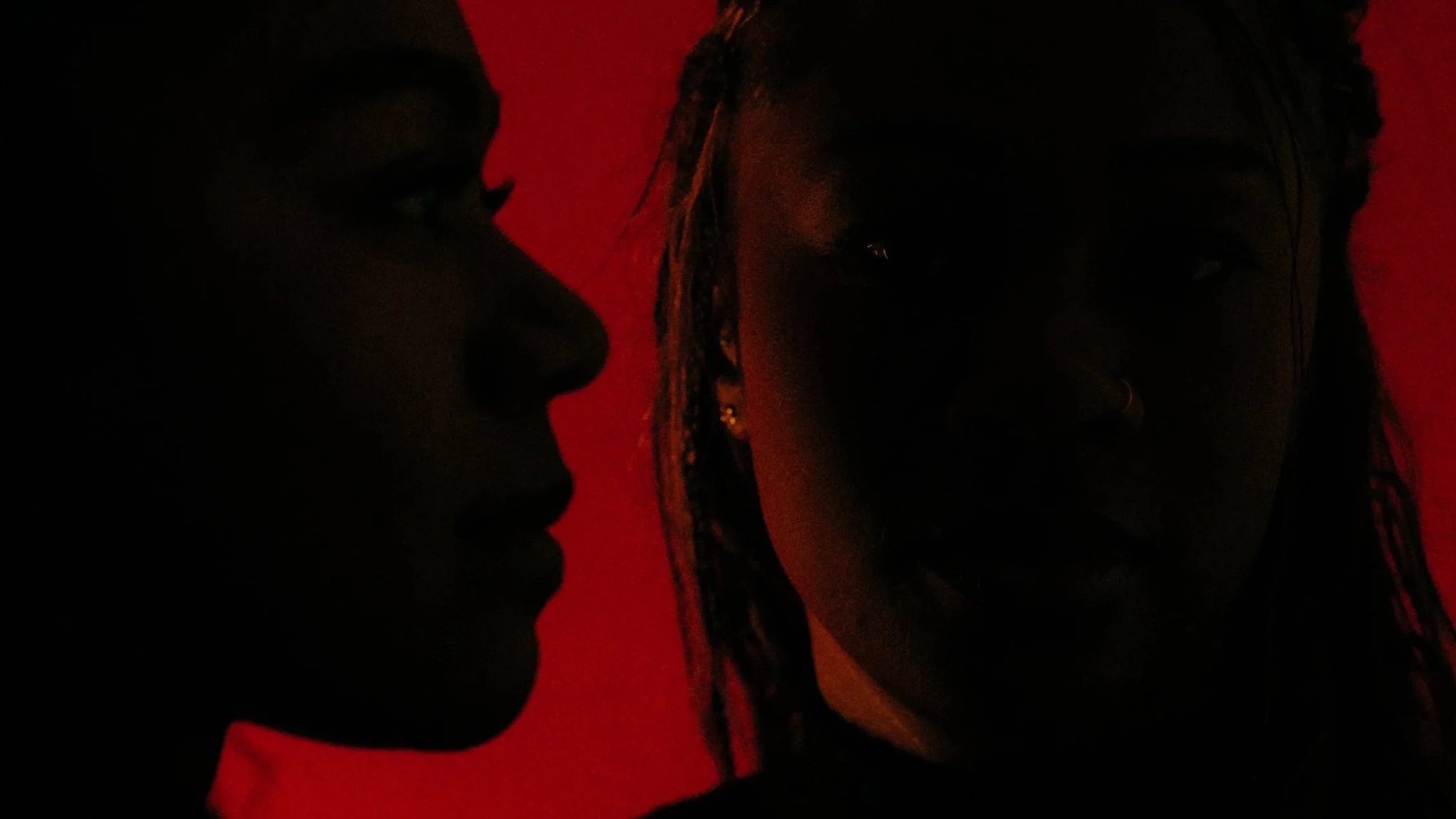 Two women with braided hair and earrings facing each other in a dark setting with a red background.