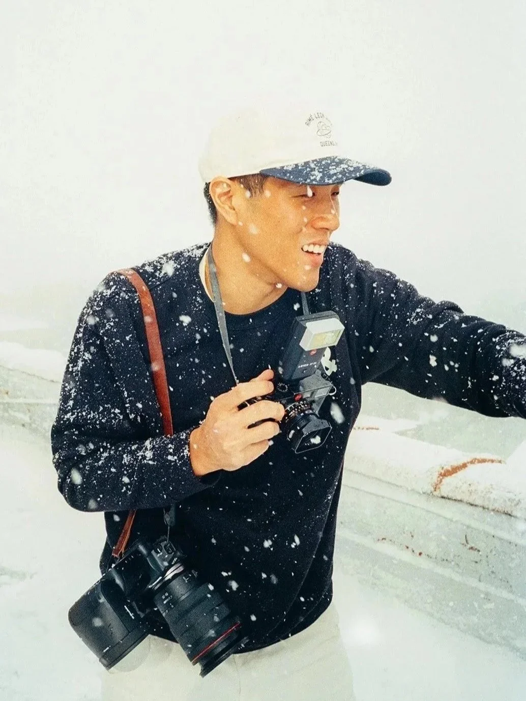 A person wearing a white cap, black sweatshirt, and beige pants is smiling and holding a camera in a snowy environment with snow falling around them.