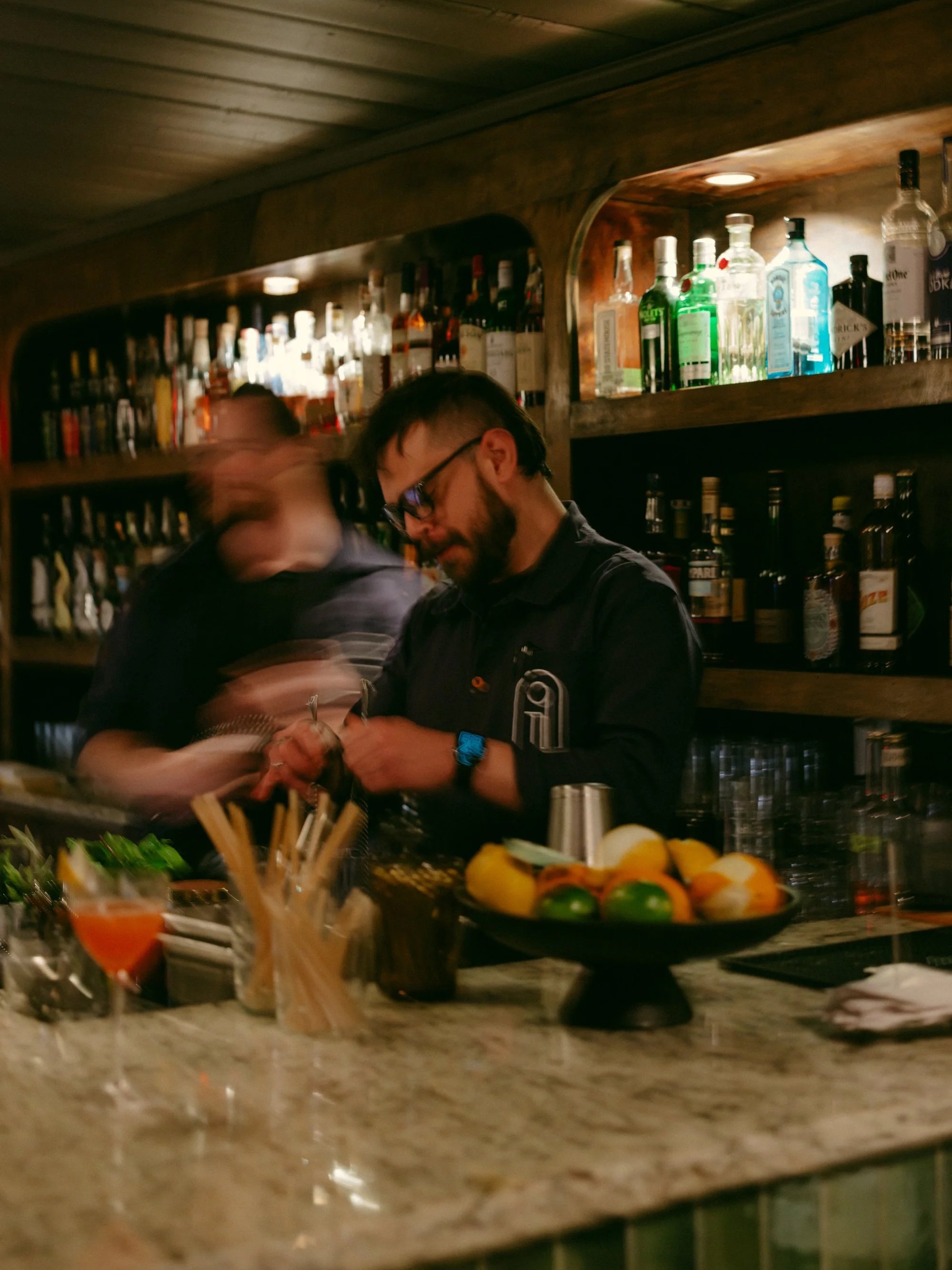 A bartender mixing drinks behind a bar counter with various bottles of alcohol on shelves in the background and fresh fruit and garnishes on the counter in the foreground.