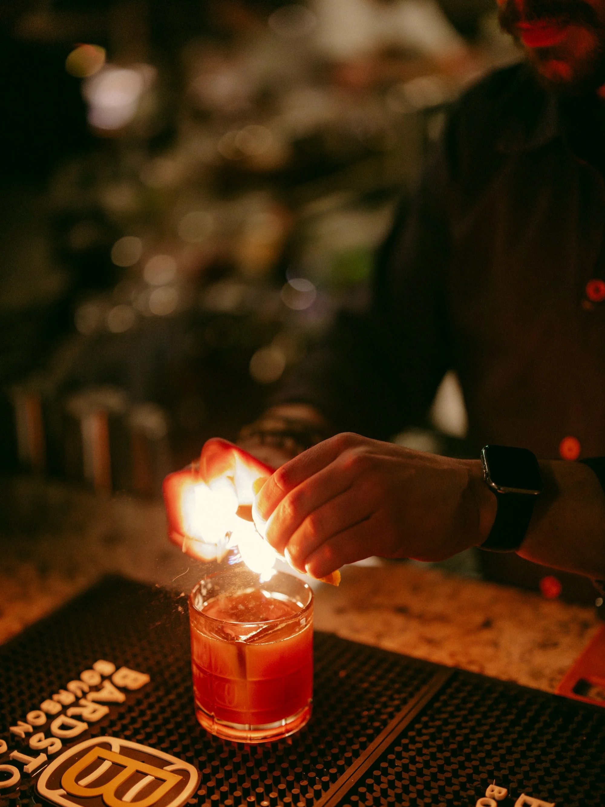 A person is holding a magazine in one hand and lighting a candle with a match in the other hand at night. There is a glass of orange-colored drink on the table in front of them, and the table has a branded mat with the logo 'DUO'. The background is d
