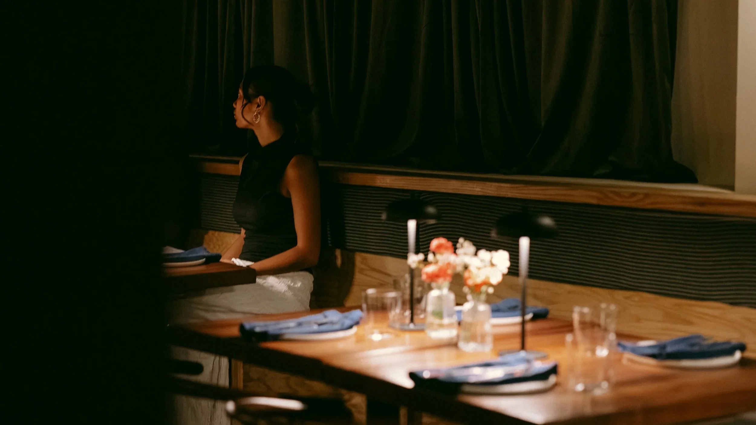 A woman with dark hair and earrings sitting at a wooden table in a dimly lit room with dark curtains in the background.
