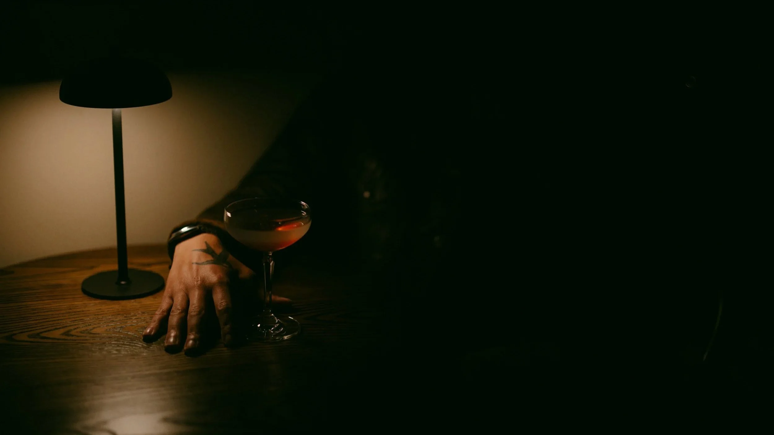 A dimly lit scene with a person's hand resting on a wooden table, holding a glass of red wine. There is a black table lamp casting a warm glow nearby.