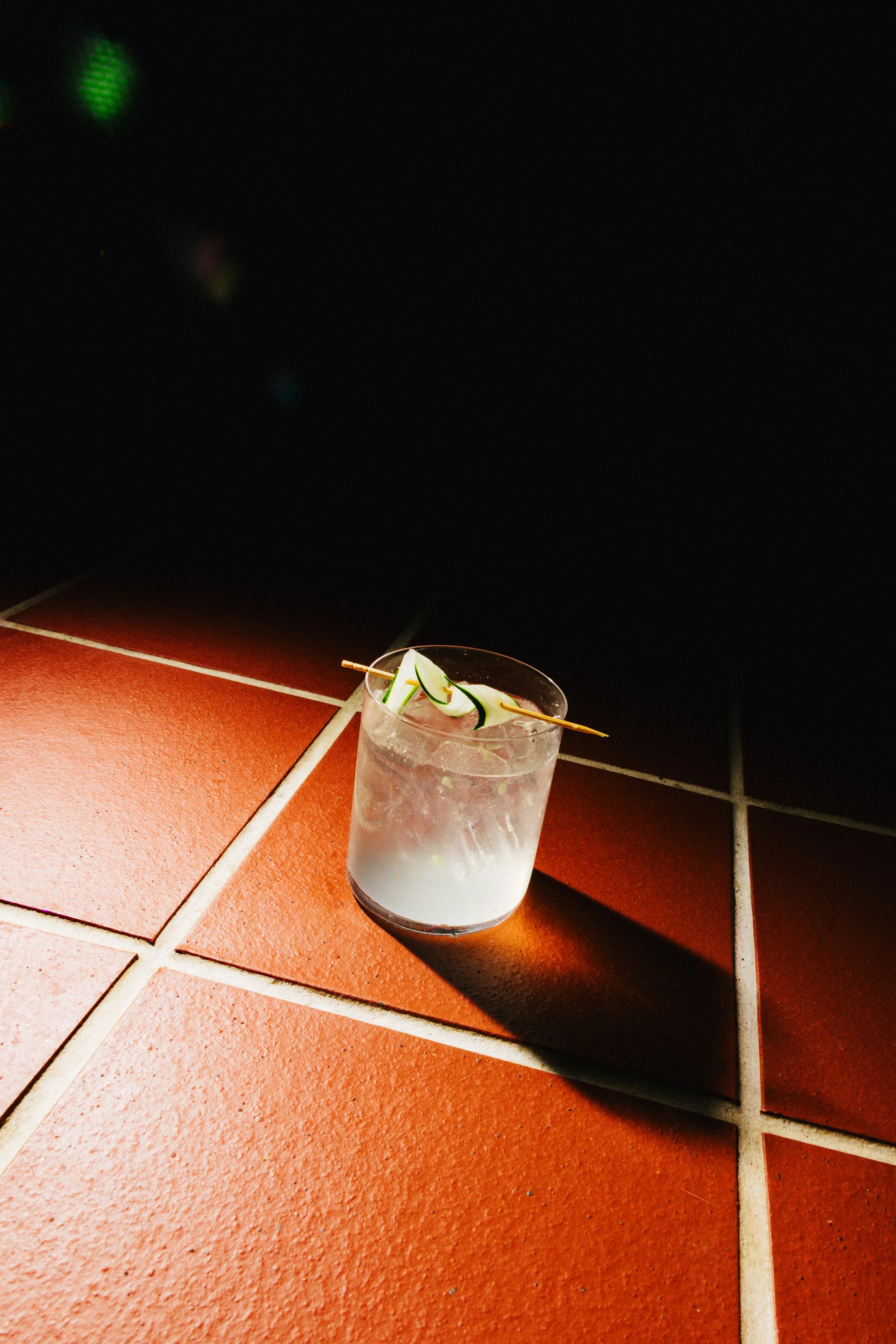 A glass with a clear drink and ice, garnished with cucumber slices, placed on a tiled floor, casting a shadow.