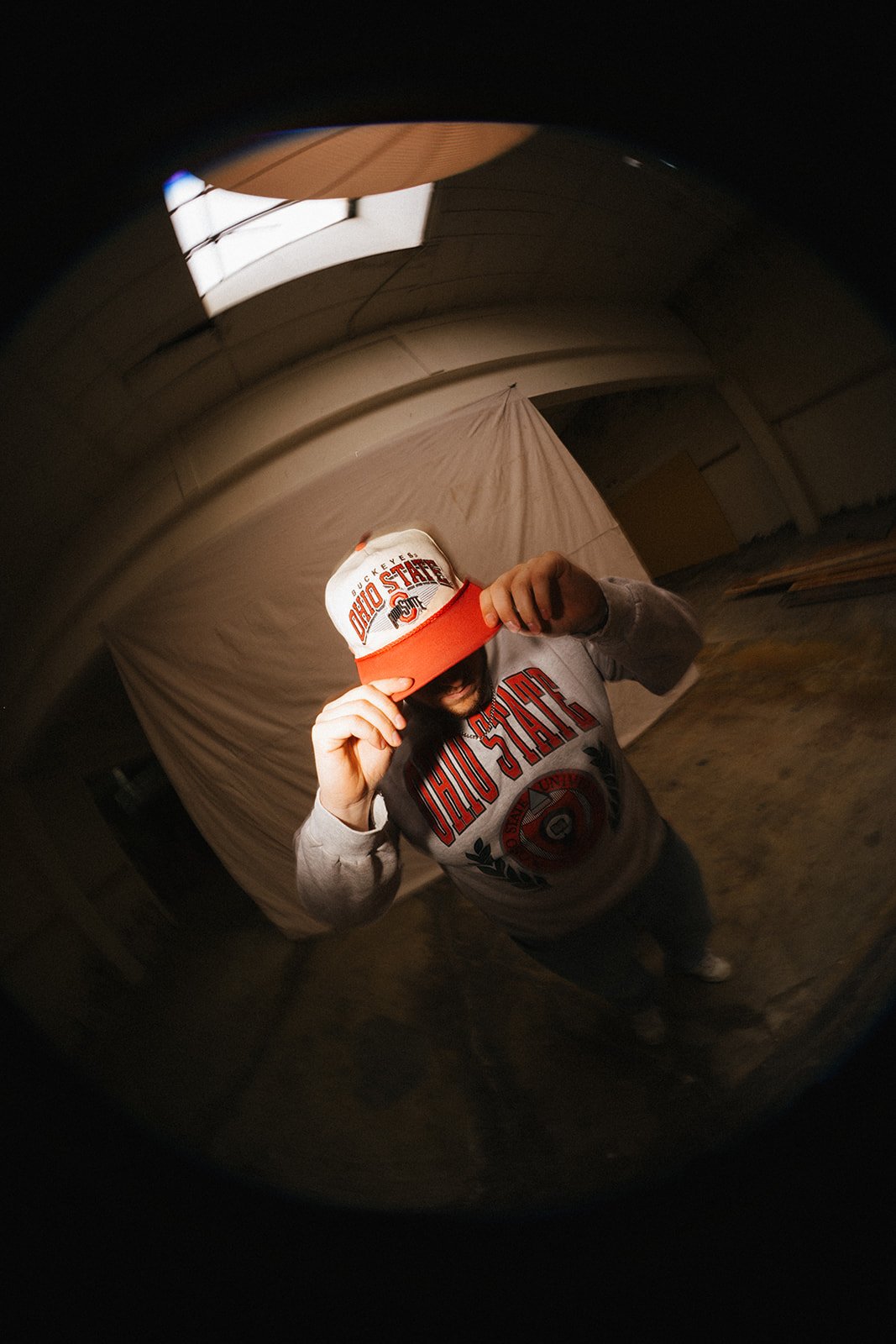 Person wearing Ohio State Buckeyes sweatshirt and cap, holding the cap's brim with one hand, standing in an indoor setting with a beige backdrop and hardwood floor, taken with a fisheye lens.