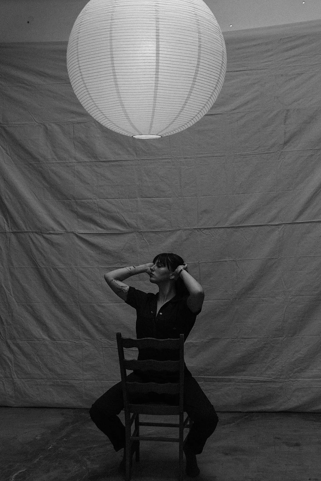 A woman sitting on a chair with her legs apart, wearing dark clothing, with her hands on her head, in front of a large textured backdrop and illuminated by a large round paper lantern hanging from the ceiling.