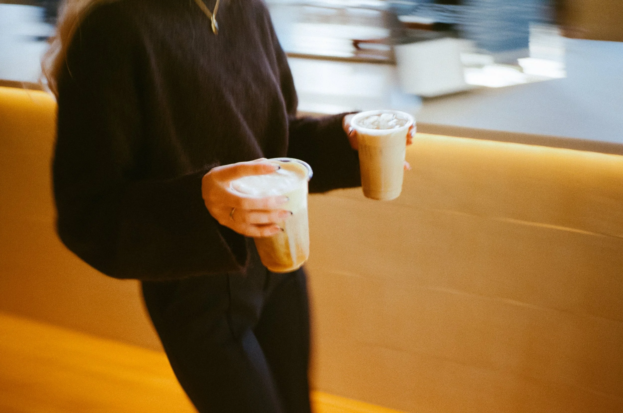 A person holding two iced coffee drinks inside a cafe with a yellow bench and a window in the background.