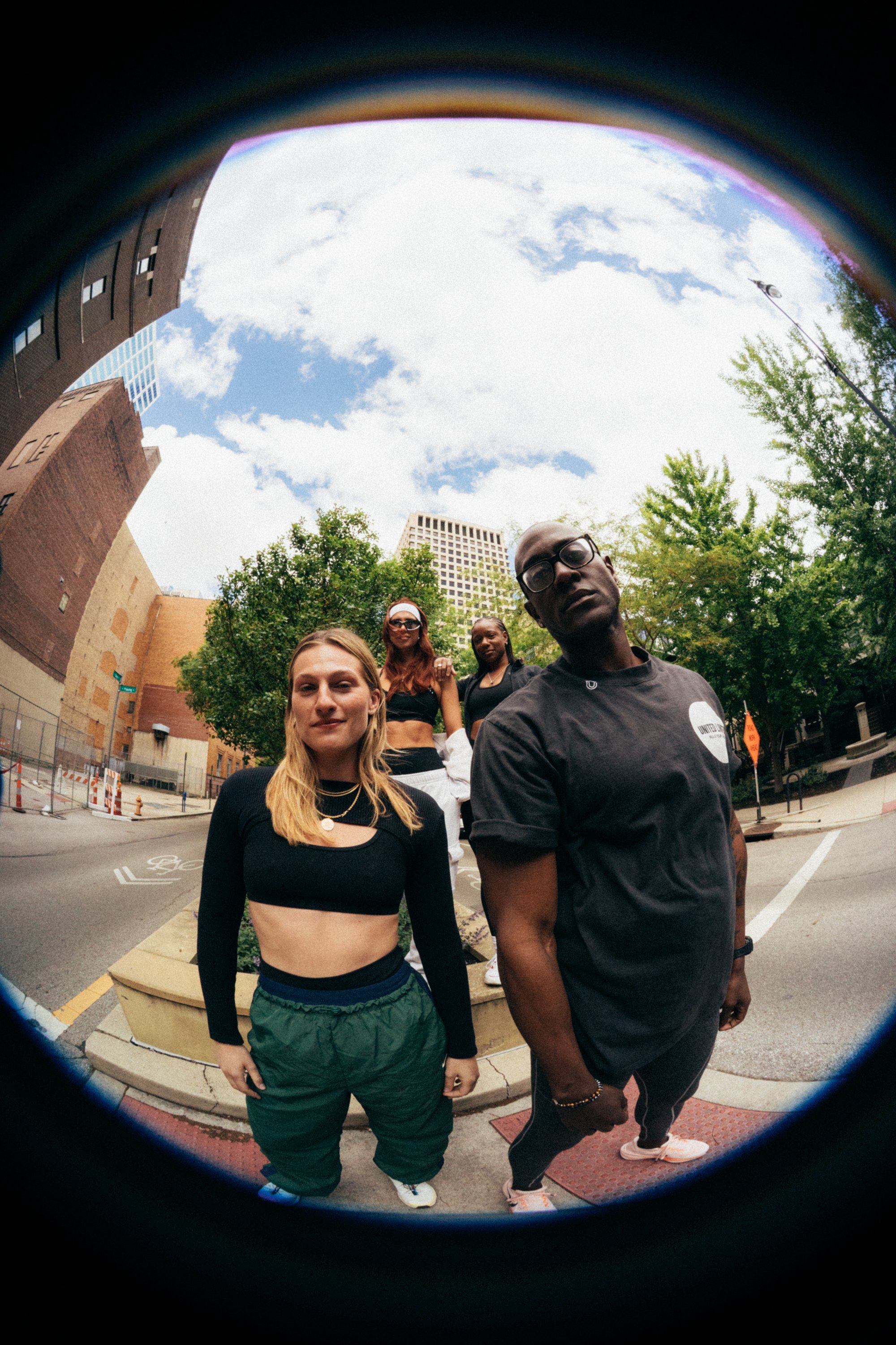Four people standing on a city sidewalk, viewed through a fisheye lens. The group includes a woman with blond hair in black workout clothes, a woman with red hair in sunglasses, a woman with braided hair in dark athletic wear, and a man with glasses 