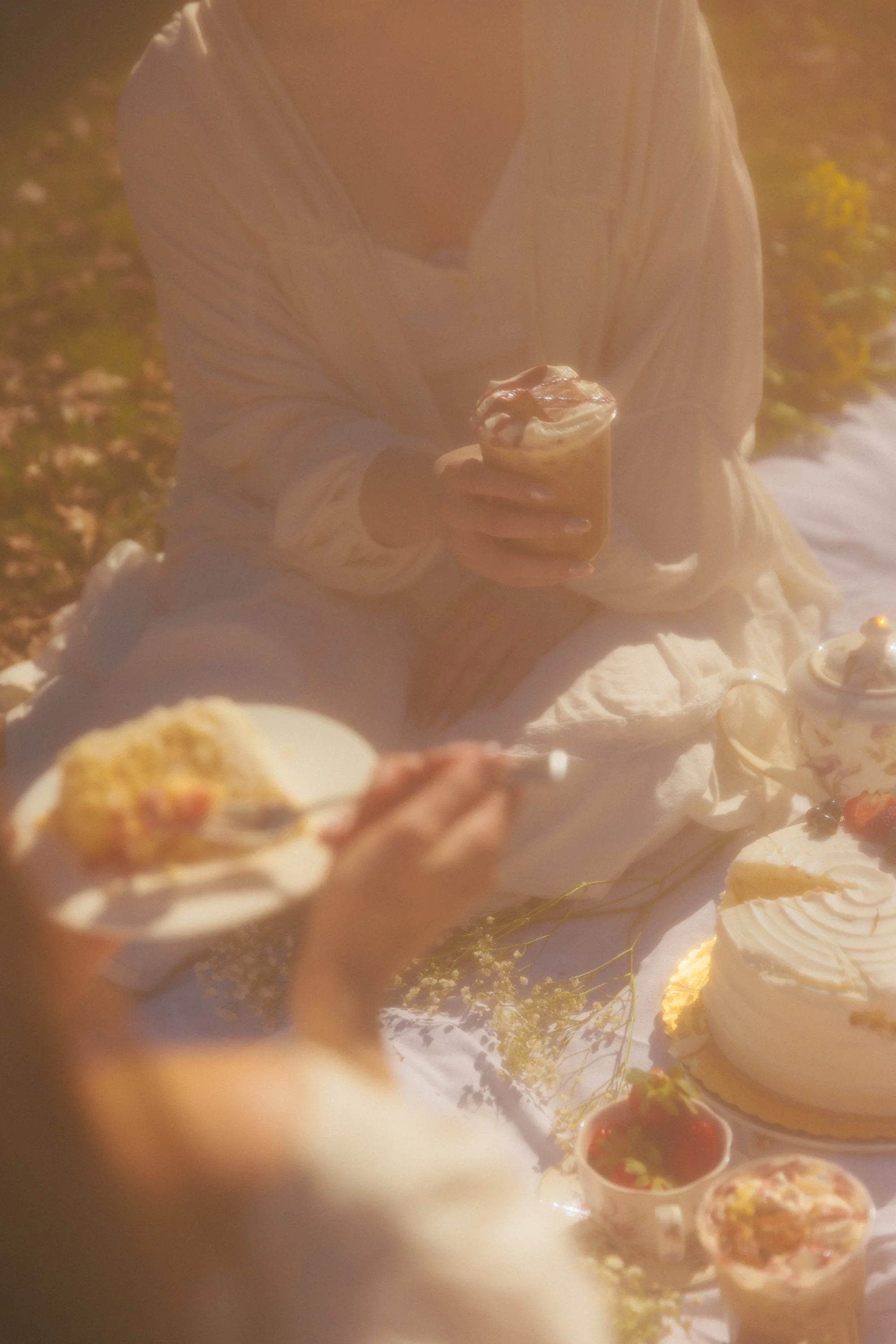 Person sitting outdoors holding a dessert in a glass, surrounded by other desserts and small bowls of sauce, with a blurred background of autumn leaves and flowers.