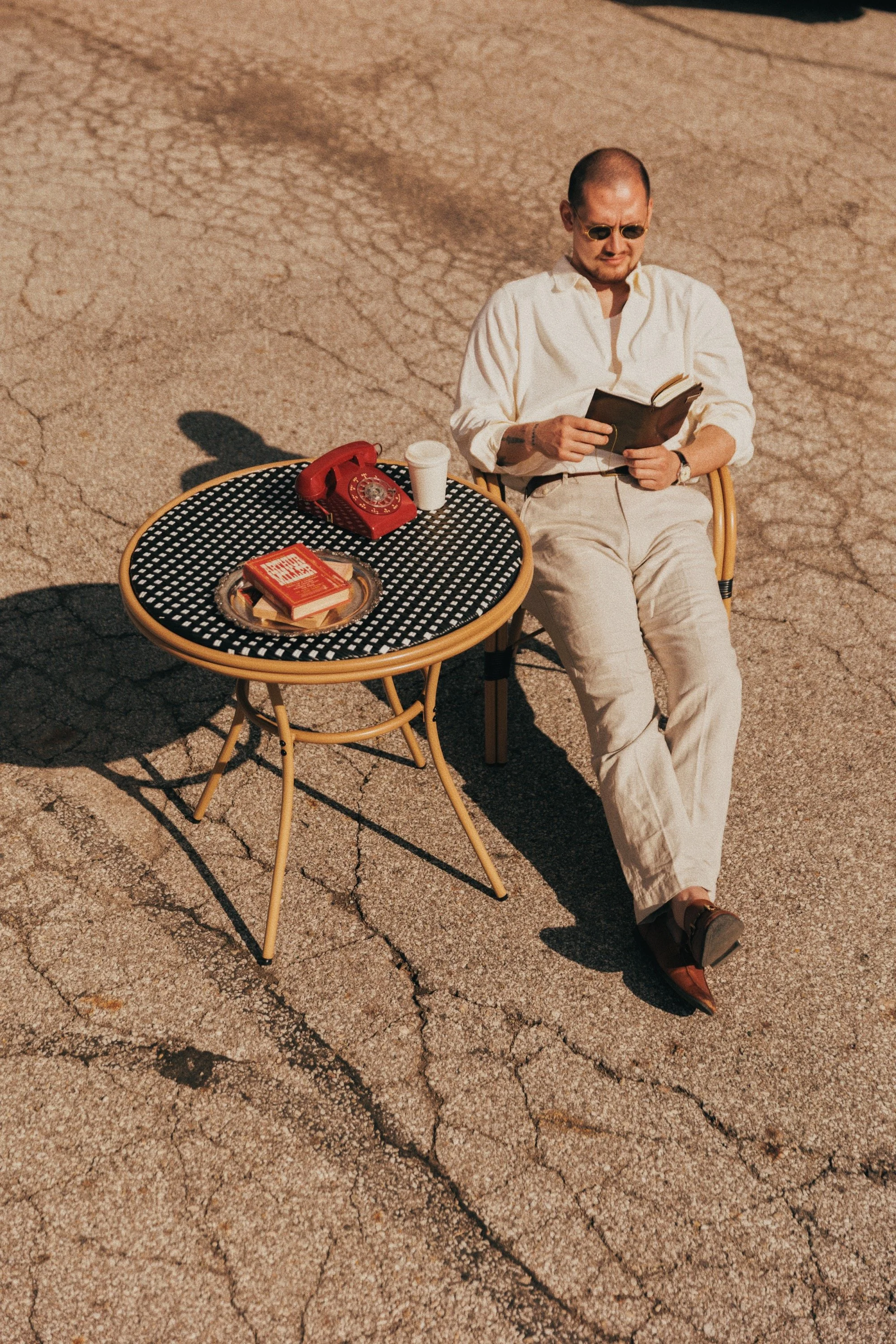 A man sitting in a chair on cracked pavement, wearing a white shirt and beige pants, reading a book. A small round table next to him has a red rotary phone, a white cup, and a tray with a book. The scene appears to be outdoors with a dry ground.