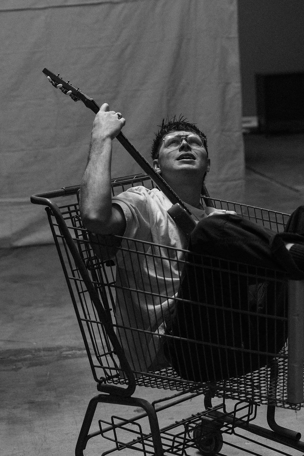 A young man with glasses and a white shirt lying in a shopping cart, holding a guitar, looking upward with a hopeful expression.