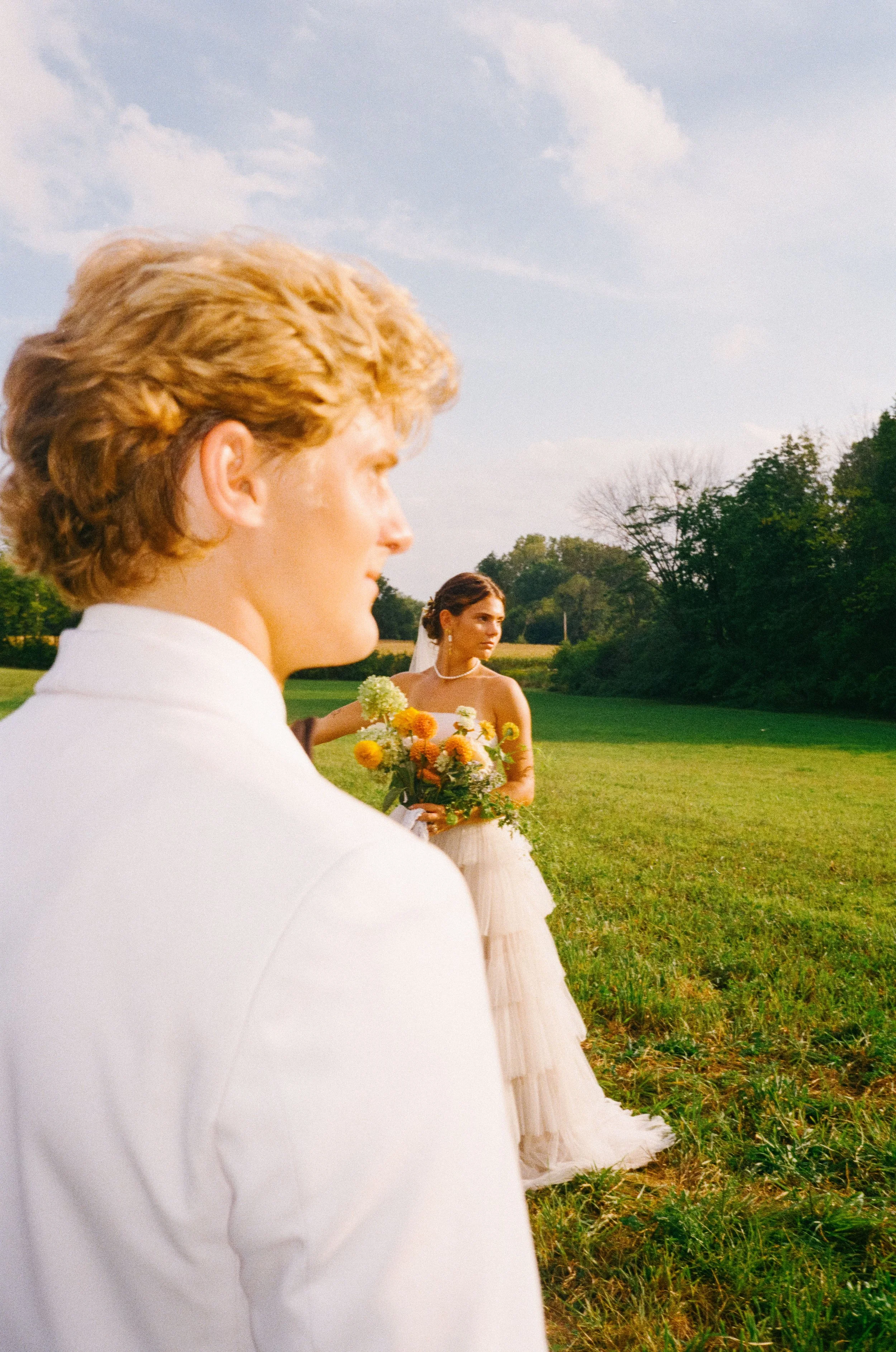 A wedding scene with a man in a white suit in the foreground and a bride holding a bouquet of flowers in the background, outdoors on a grassy field under a partly cloudy sky.