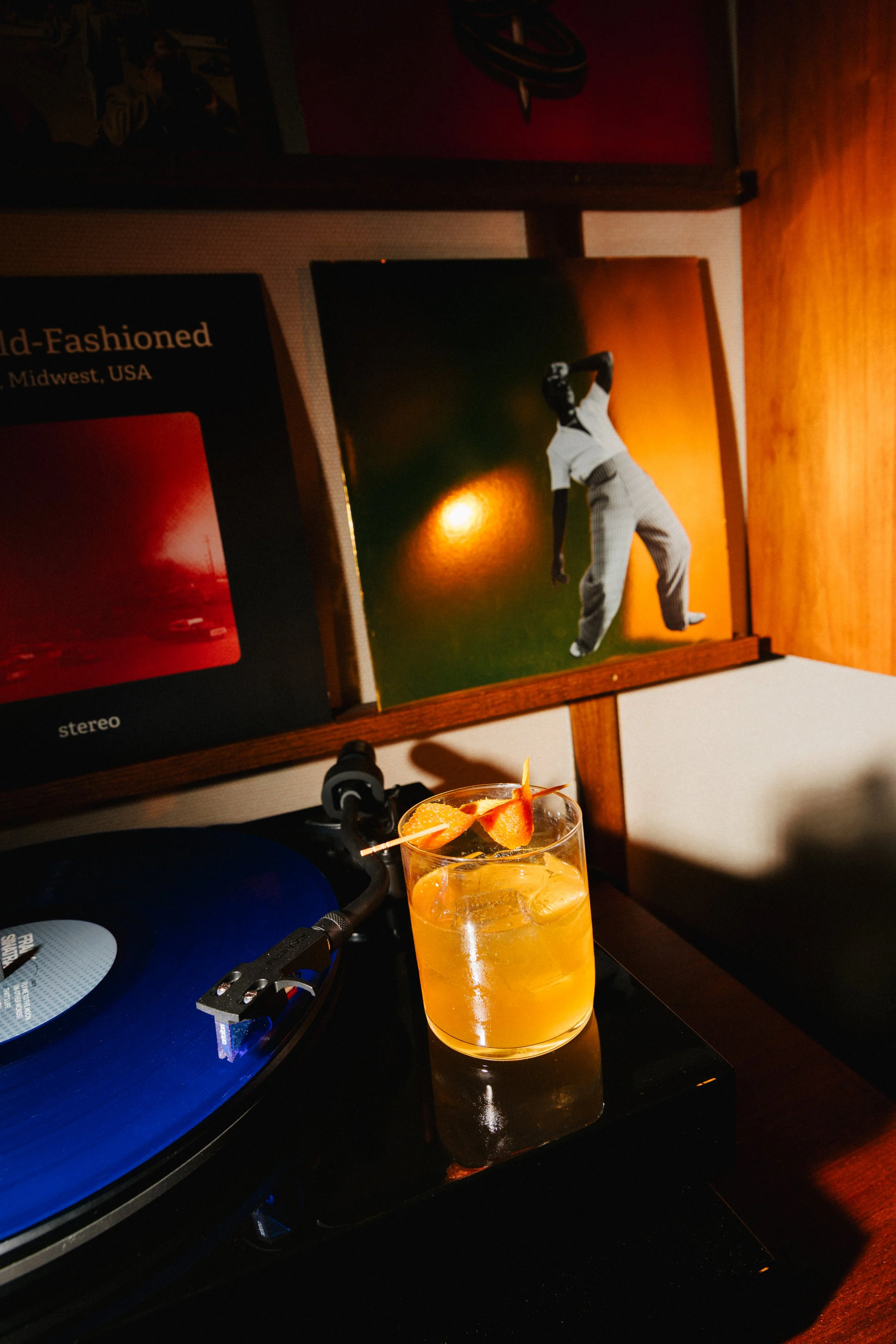 A turntable with a blue vinyl record, a glass of orange-colored drink with fruit garnishes, and two vinyl record album covers propped up against a wall, one showing a person in a white shirt and checkered pants in a dance pose, the other partially vi