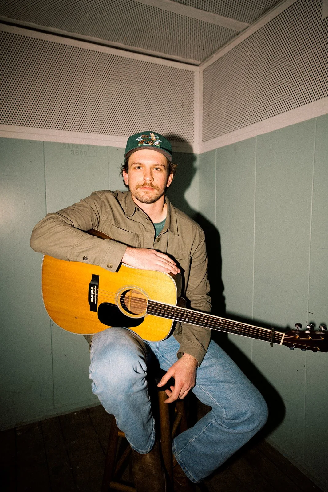 Man sitting on a wooden stool, holding an acoustic guitar, wearing a green cap, beige jacket, and jeans, in a room with gray walls and ceiling.