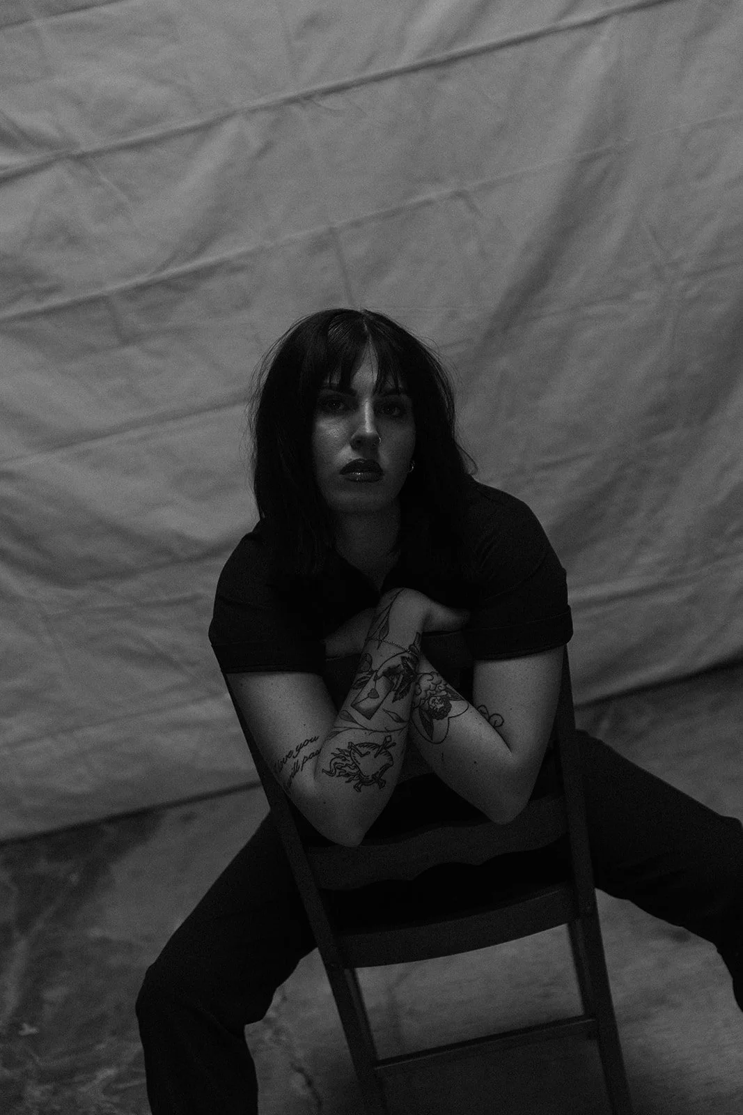 A black and white photo of a woman with dark hair and tattoos on her arms, sitting on a chair with her hands clasped together in front of her, against a textured background.
