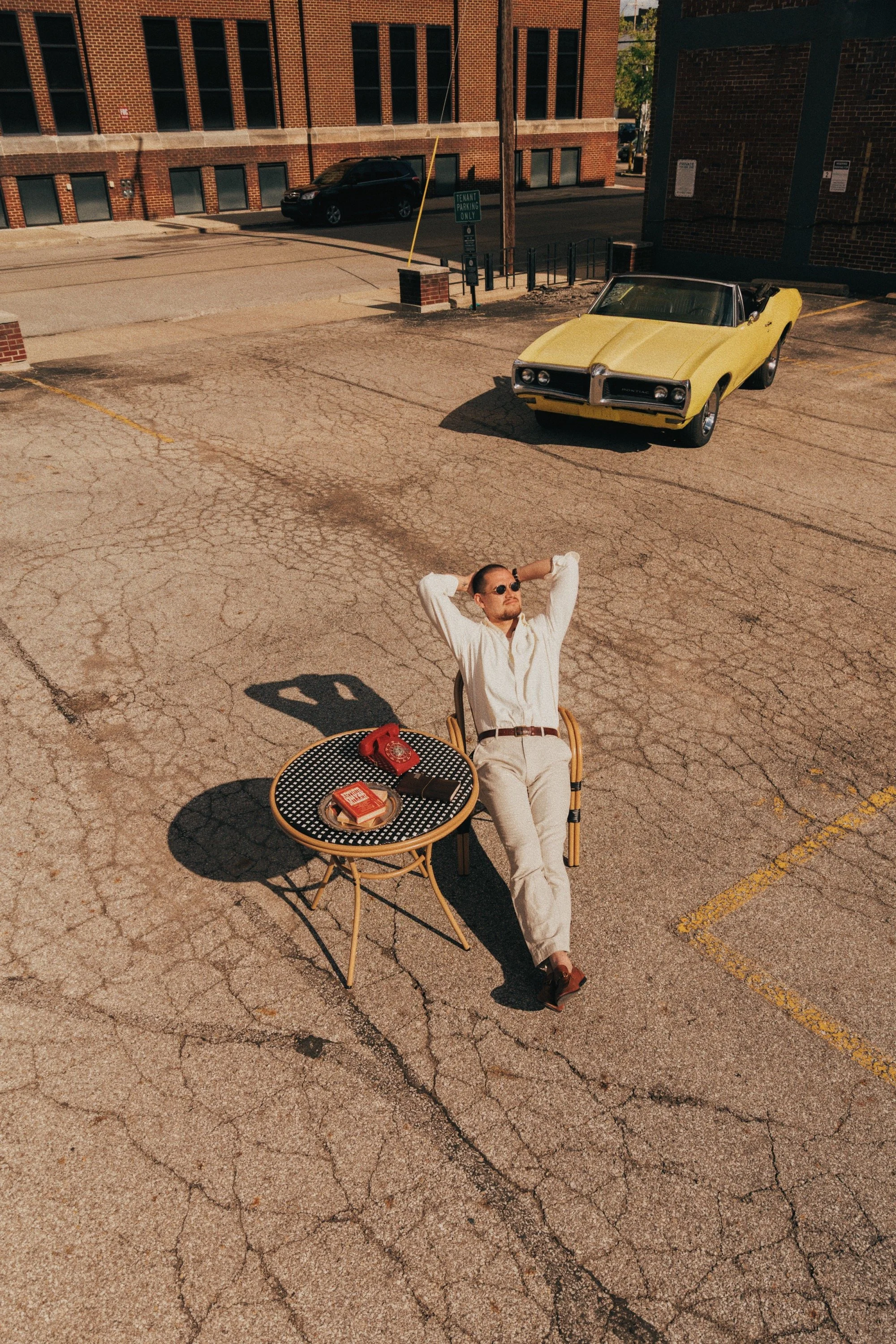 A man wearing a white shirt and beige pants is sitting on a chair in an outdoor parking lot, relaxing with his hands behind his head. There is a small round table in front of him with a red rotary phone, a pack of cigarettes, and a lighter. A yellow 