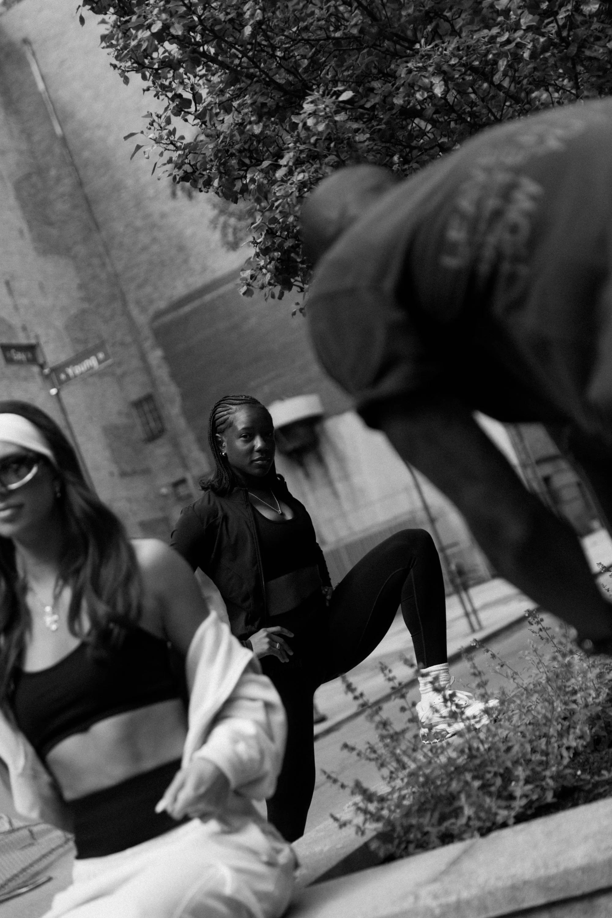 Black and white photo of three women sitting outdoors near a wall and trees. One woman in the background looks at the camera, while the other two women are partially visible and out of focus.