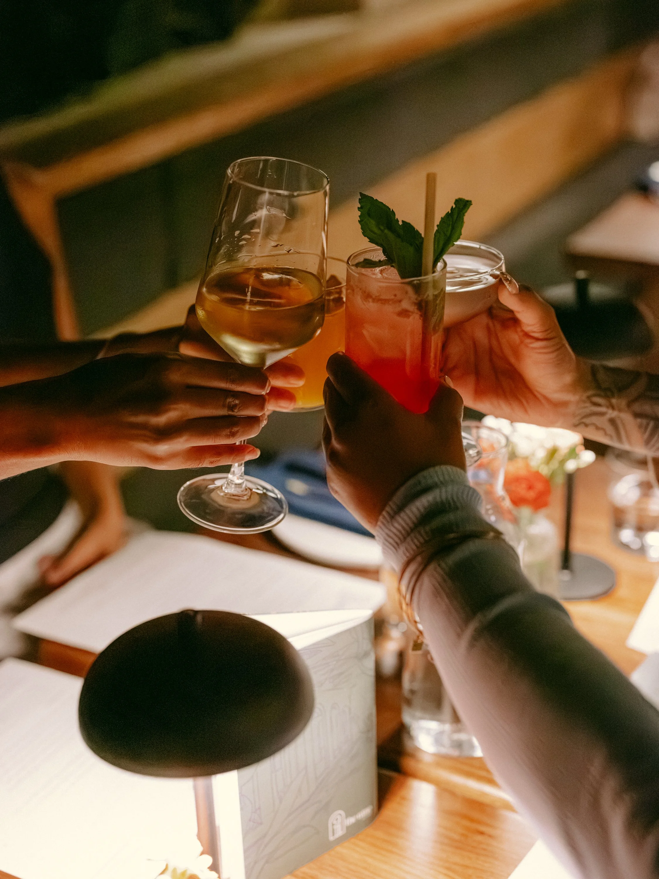 People raising glasses in a toast, one with white wine, one with a pink cocktail garnished with mint leaves, in a cozy indoor setting.