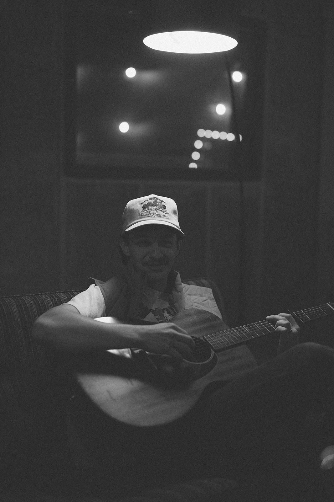 A person wearing glasses, a white cap, and a vest playing an acoustic guitar in a dimly lit room.