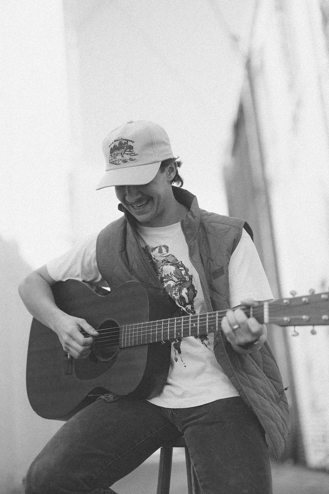 A young man sitting on a stool playing an acoustic guitar, smiling, wearing a cap, t-shirt, and vest, in black and white.