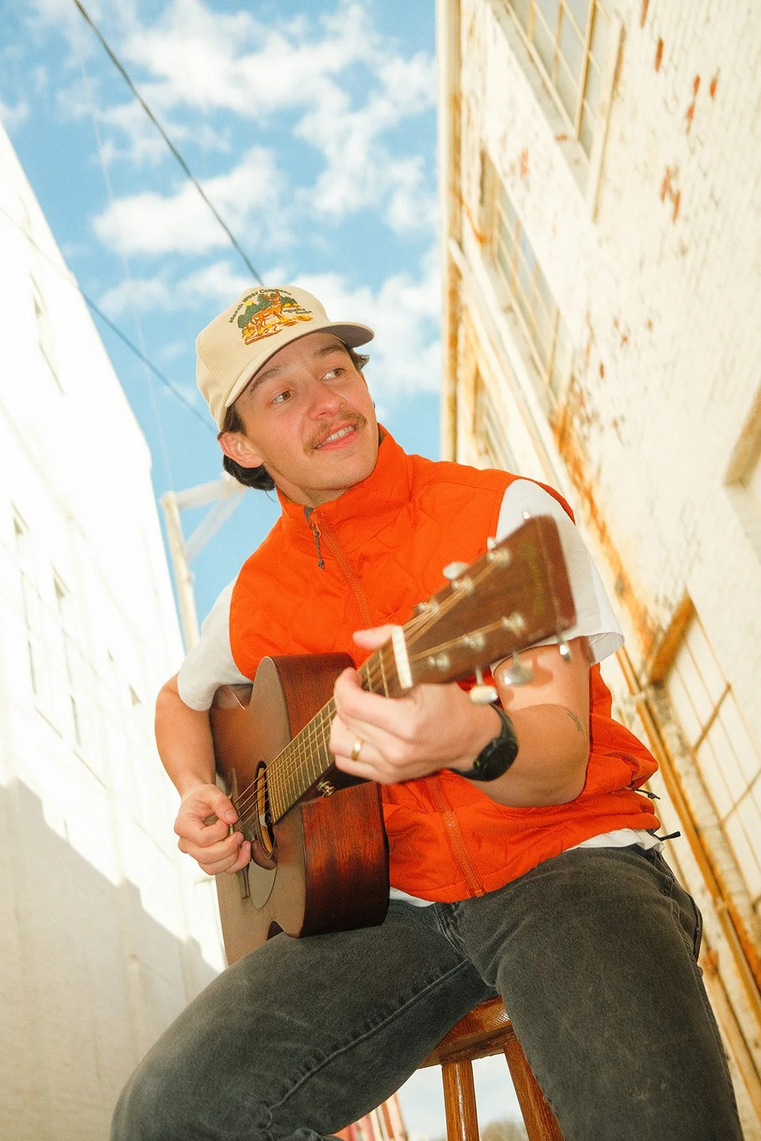 A young man playing an acoustic guitar outdoors, sitting on a wooden stool, wearing an orange vest and a cap with a colorful logo, smiling and looking off to the side with a blue sky with clouds in the background.
