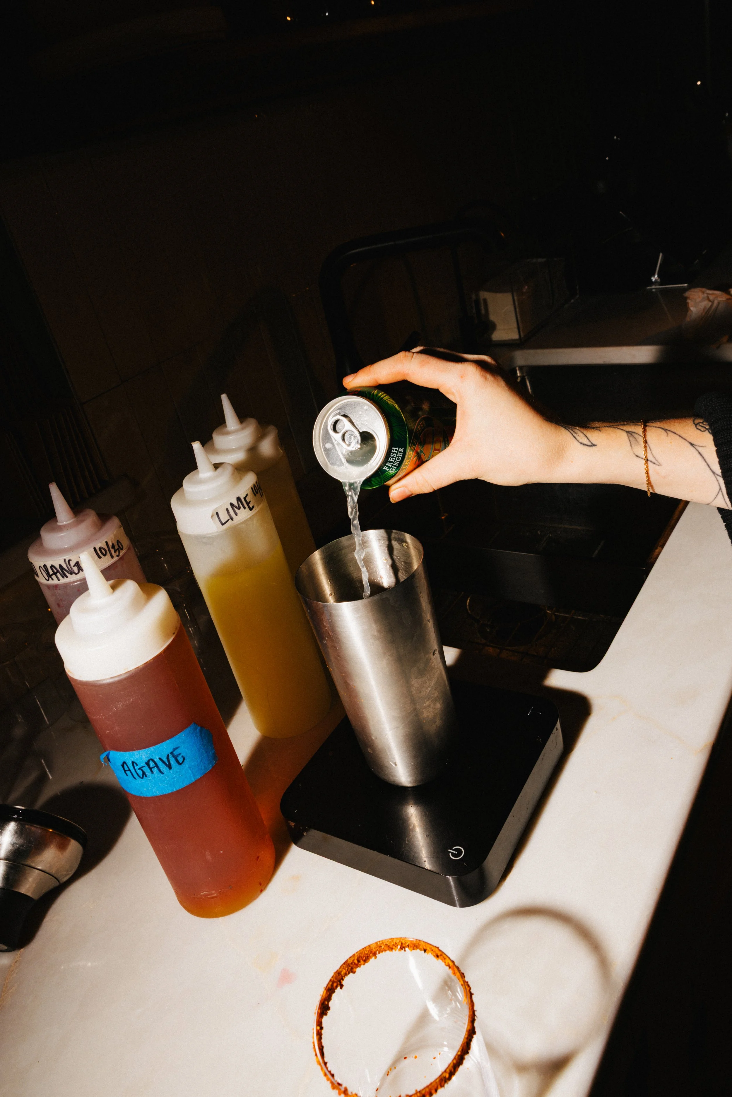 Person pouring clear liquid from a can into a stainless steel shaker on a kitchen counter, with condiment bottles labeled for different ingredients nearby.