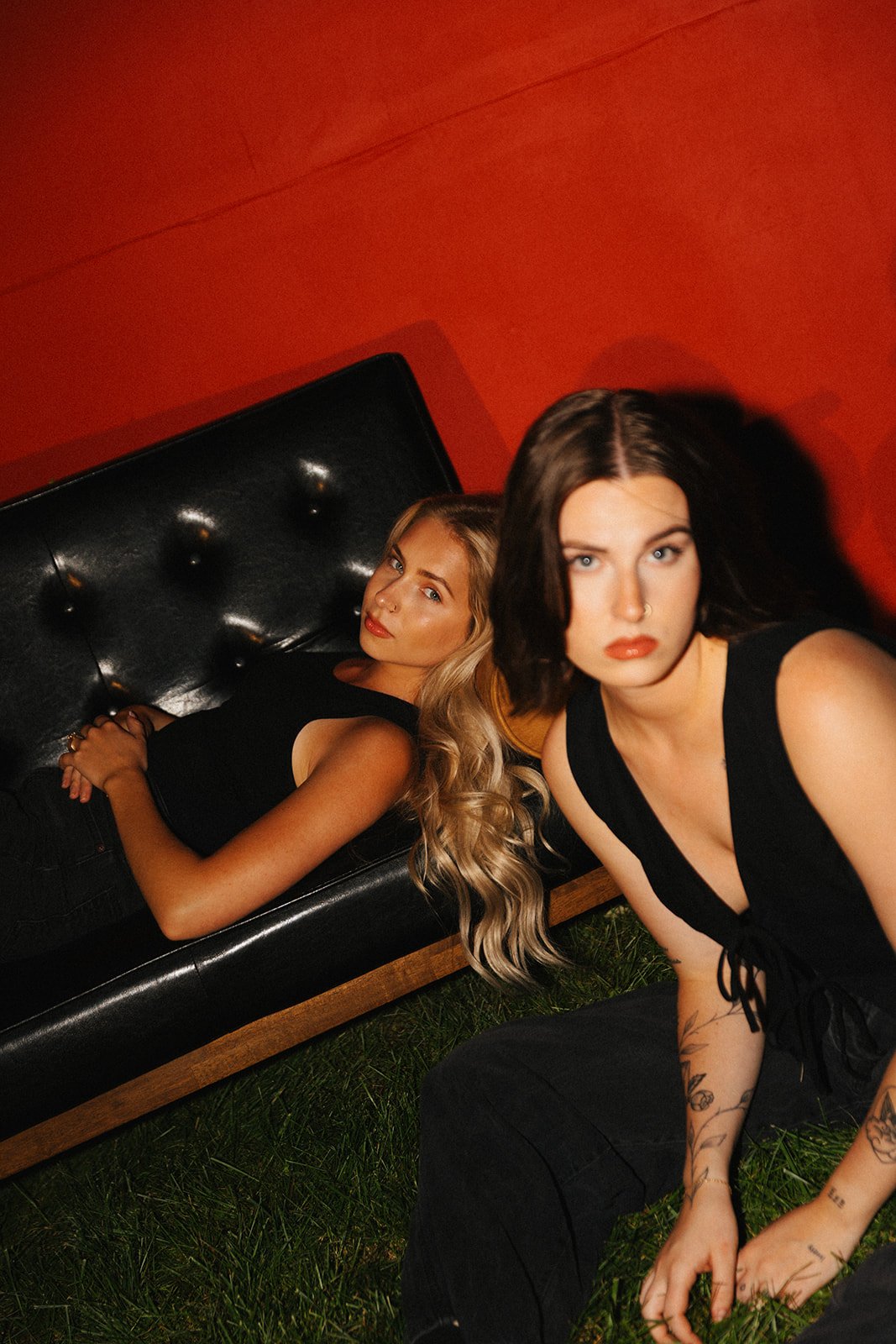 Two women, one lying on a black leather couch and the other kneeling on grass in front of her, pose against an orange wall. Both women are wearing black outfits.