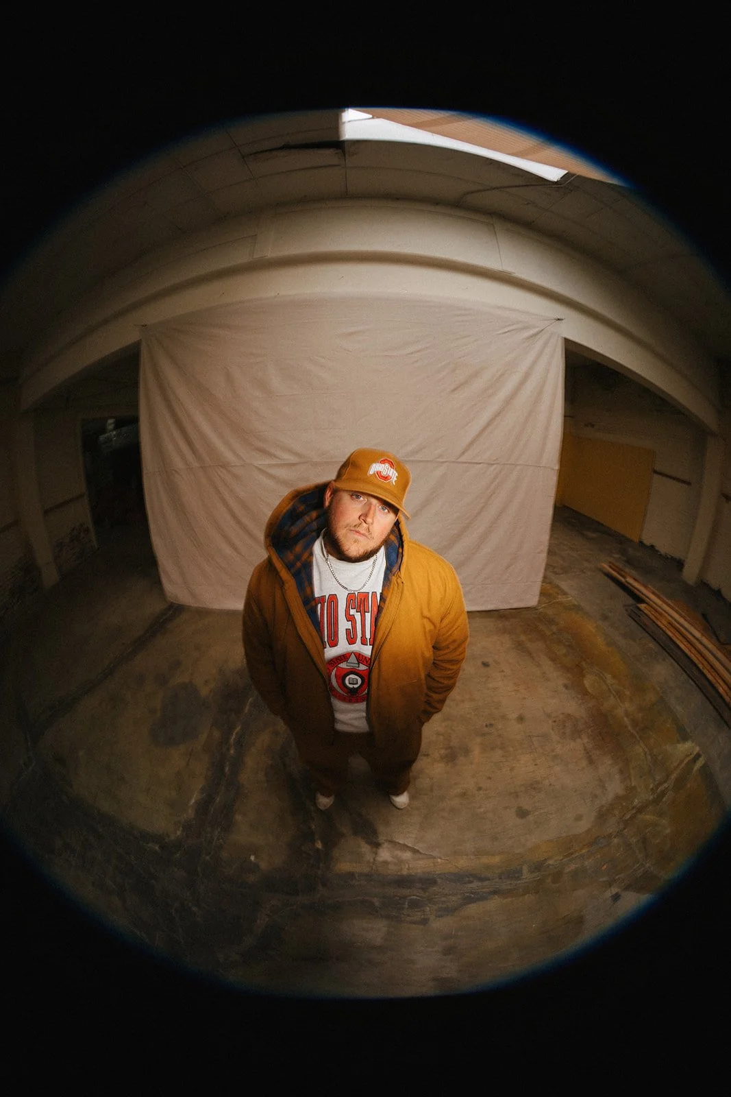 A young man wearing a Ohio State hoodie, tan jacket, and matching cap, standing with hands in pockets in an indoor space with a concrete floor and a hanging white backdrop, seen through a fisheye lens.