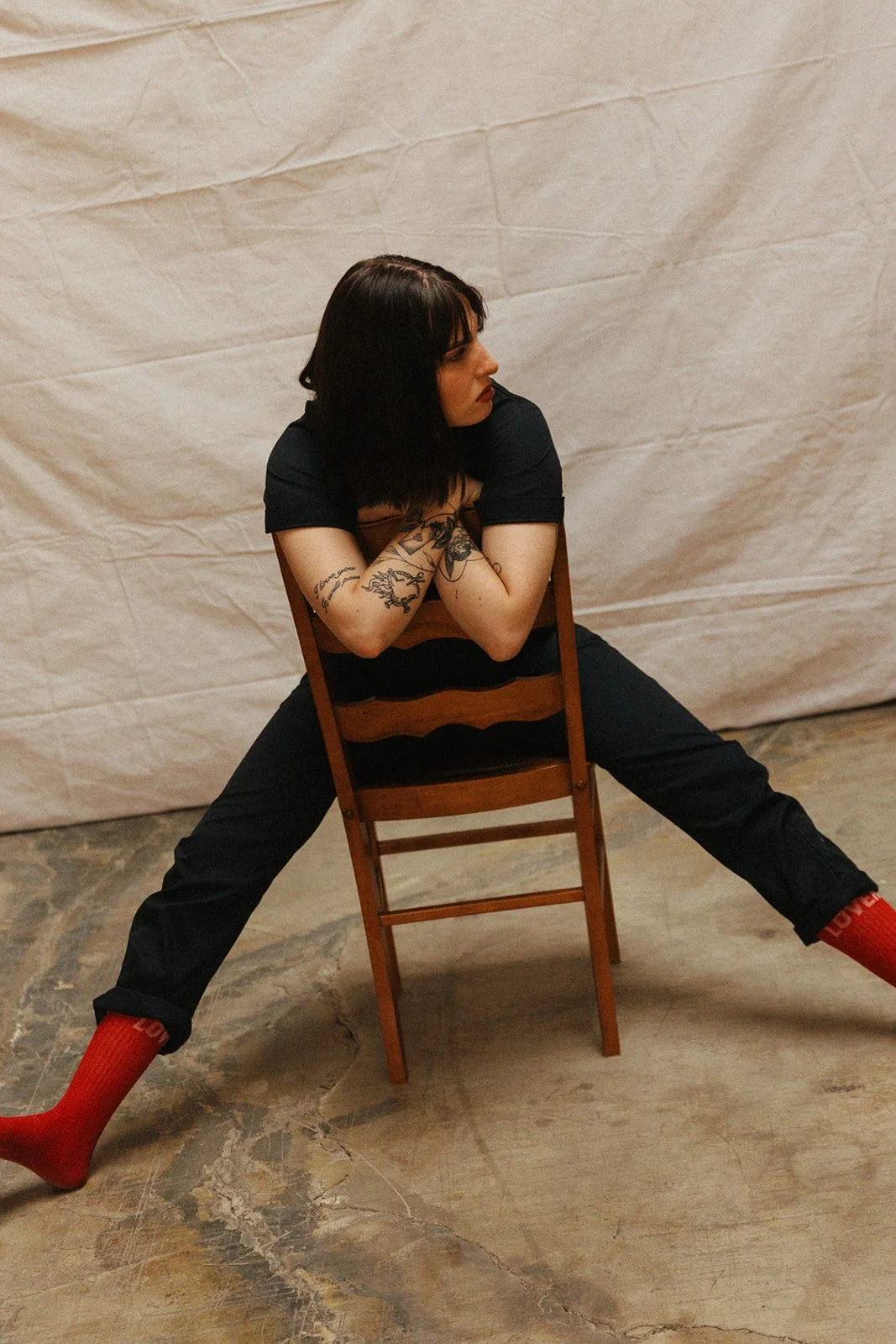 A woman with dark hair, tattoos on her arms, wearing a black shirt, black pants, and red socks, sitting backwards on a wooden chair with her legs spread apart and arms crossed, against a plain background.