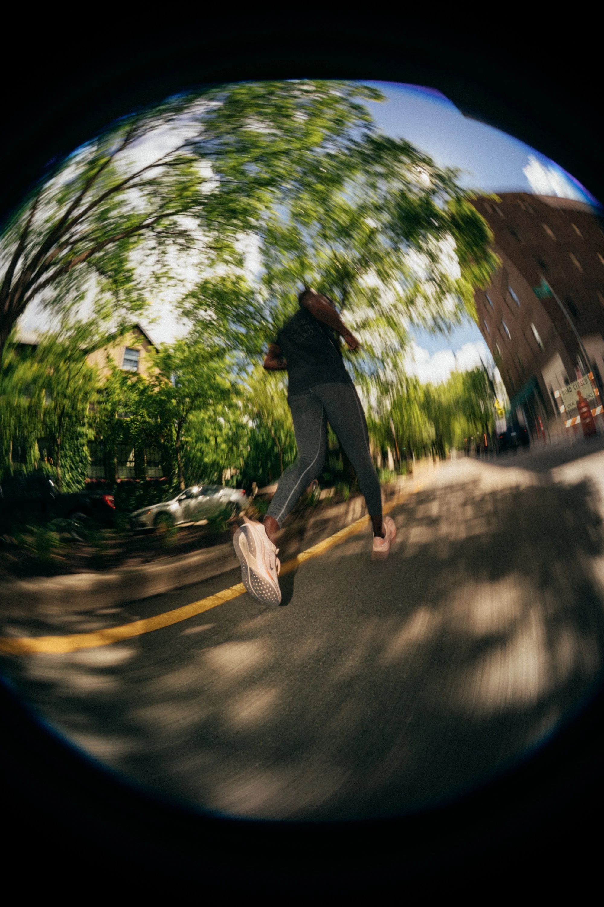 A person running on the street, captured with a fisheye lens that creates a circular distortion, with trees and buildings in the background and parked cars on the side of the road.