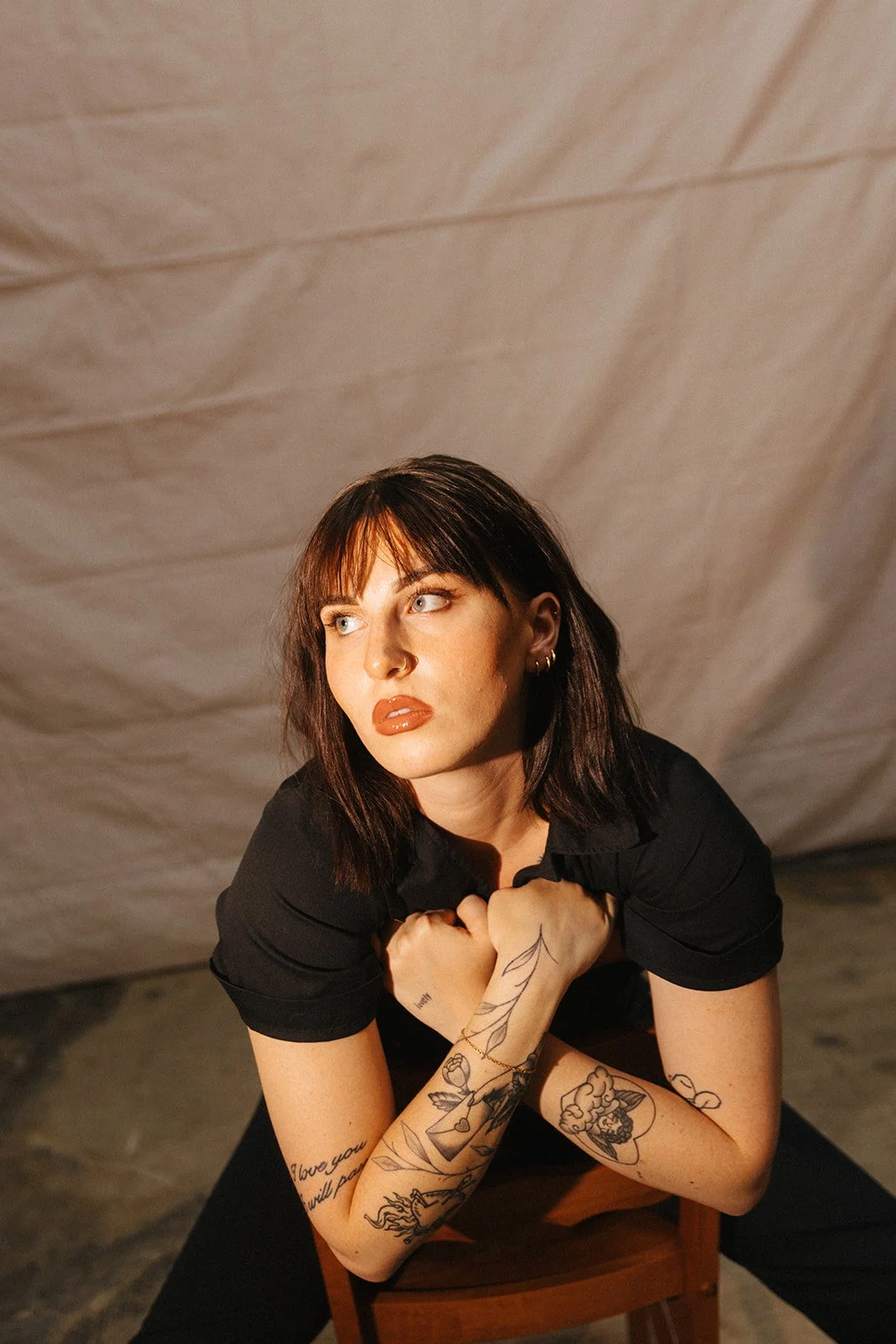 A woman with dark hair and tattoos on her arms, wearing a black shirt, sitting on a chair with her arms crossed over her chest, looking to the side in a contemplative pose.