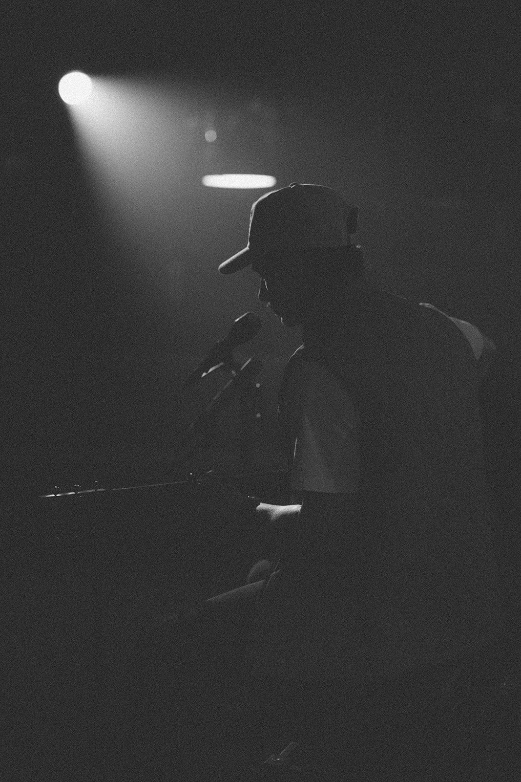 Silhouette of a person wearing a baseball cap, singing into a microphone on stage, with spotlight overhead in black and white.