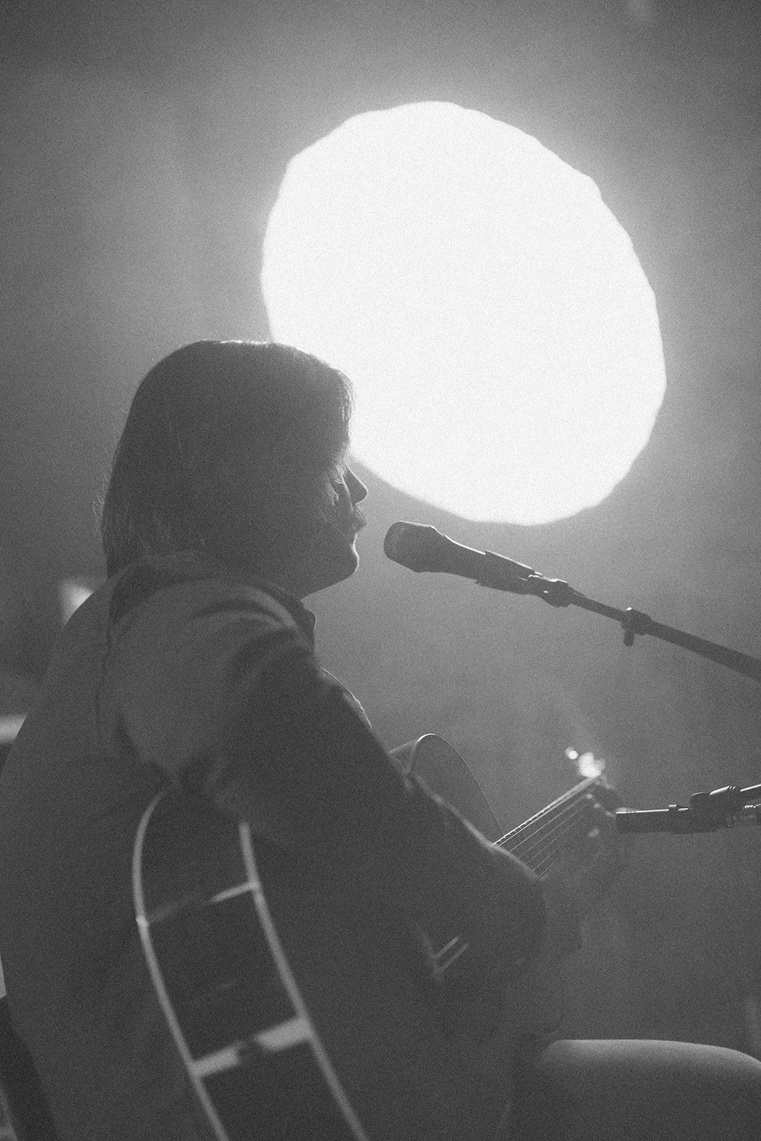 A person playing guitar and singing into a microphone, in silhouette against a bright round light in the background.
