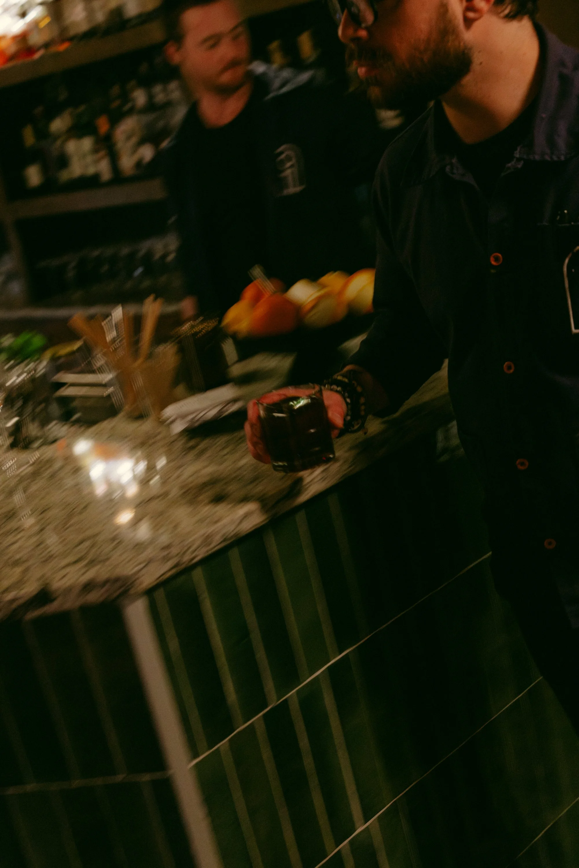 Two men are in a bar or restaurant. One man with glasses and a beard is holding a drink, the other man is in the background holding a plate of fruit. Shelves of bottles are visible in the background.