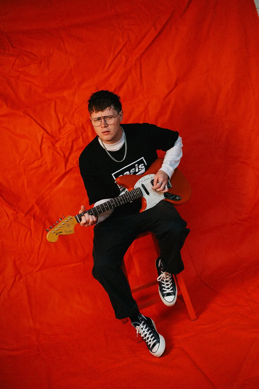 A young man wearing glasses, a black Oasis t-shirt, black pants, and black and white sneakers, sitting on a small orange stool, holding an orange electric guitar, in front of a wrinkled orange background.