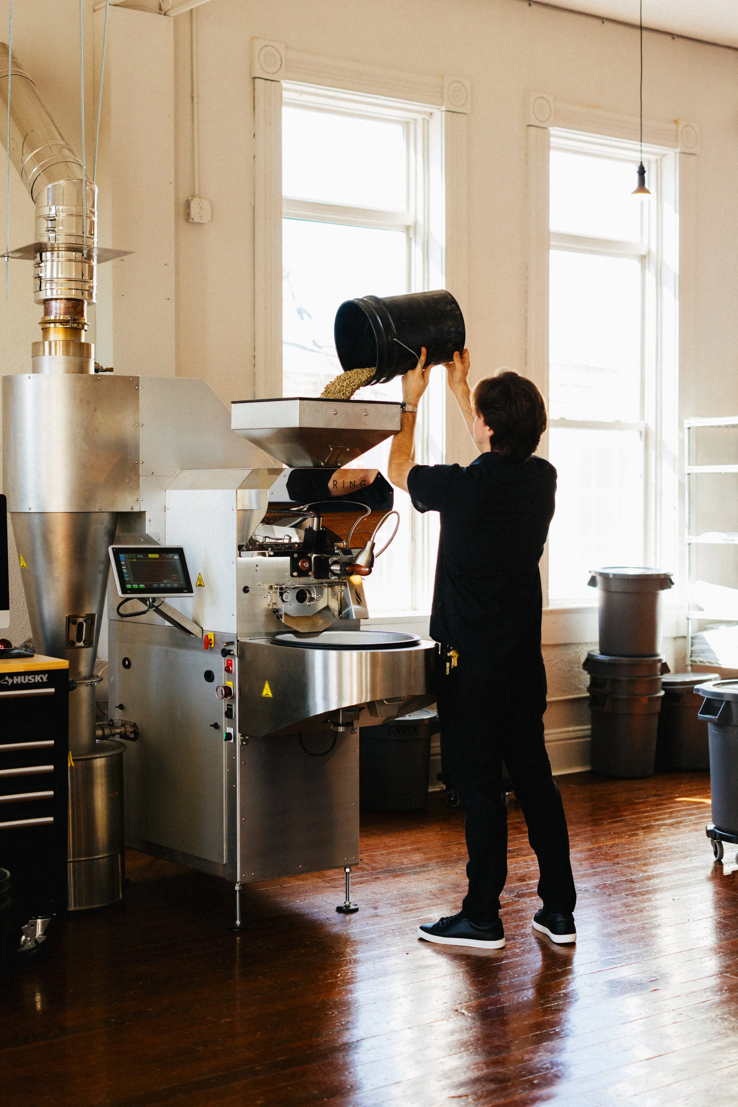 A person pouring coffee beans from a black bucket into a coffee roasting machine in a bright industrial-style room with large windows and wooden floors.