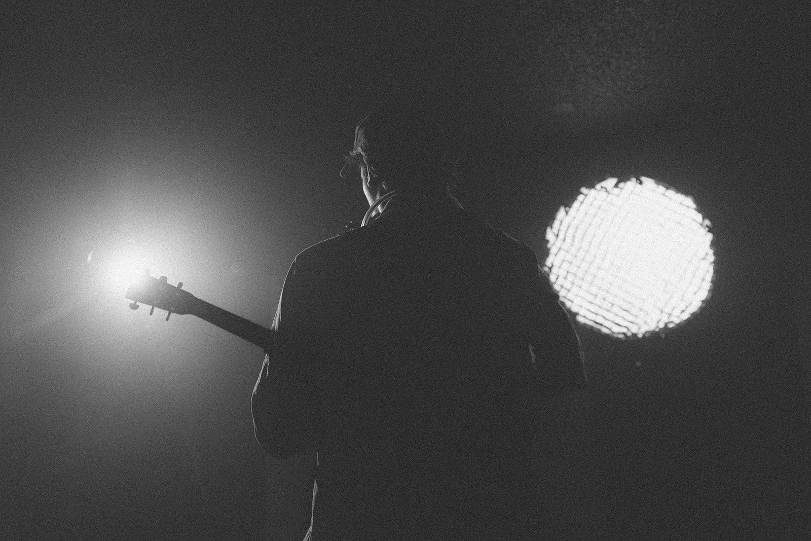 A silhouette of a person holding a guitar in front of bright stage lights, with one bright light behind them creating a contrast.