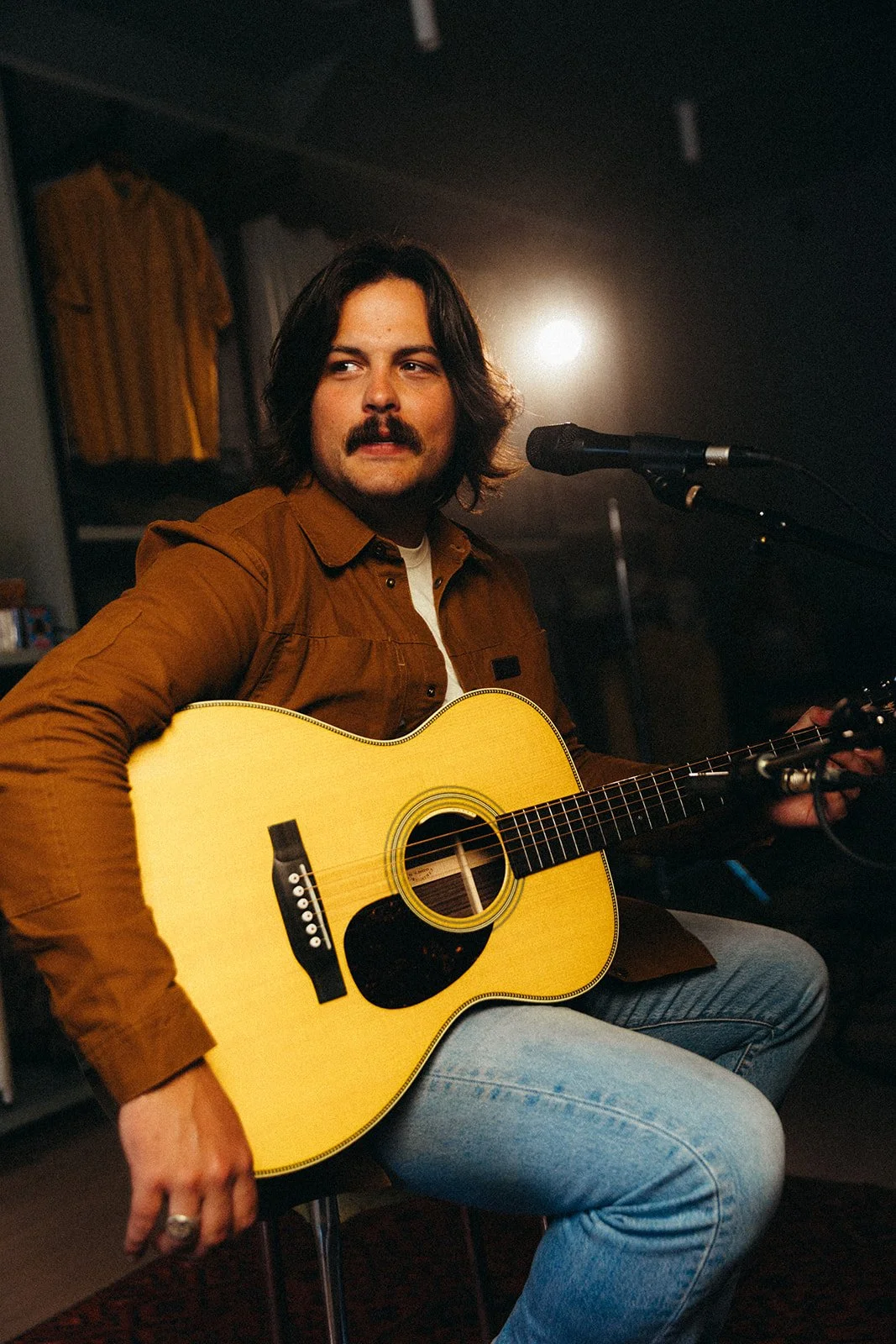 A man with dark hair, a mustache, wearing a brown jacket and blue jeans, sitting and playing an acoustic guitar, with a microphone in front of him, in a dimly lit room.